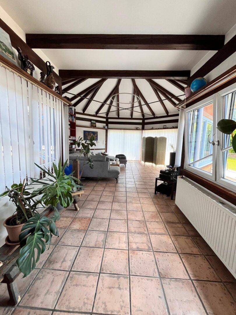 Geräumiges Wohnzimmer mit Terrakottafliesen, hohen Decken und großen Fenstern, umgeben von Pflanzen.