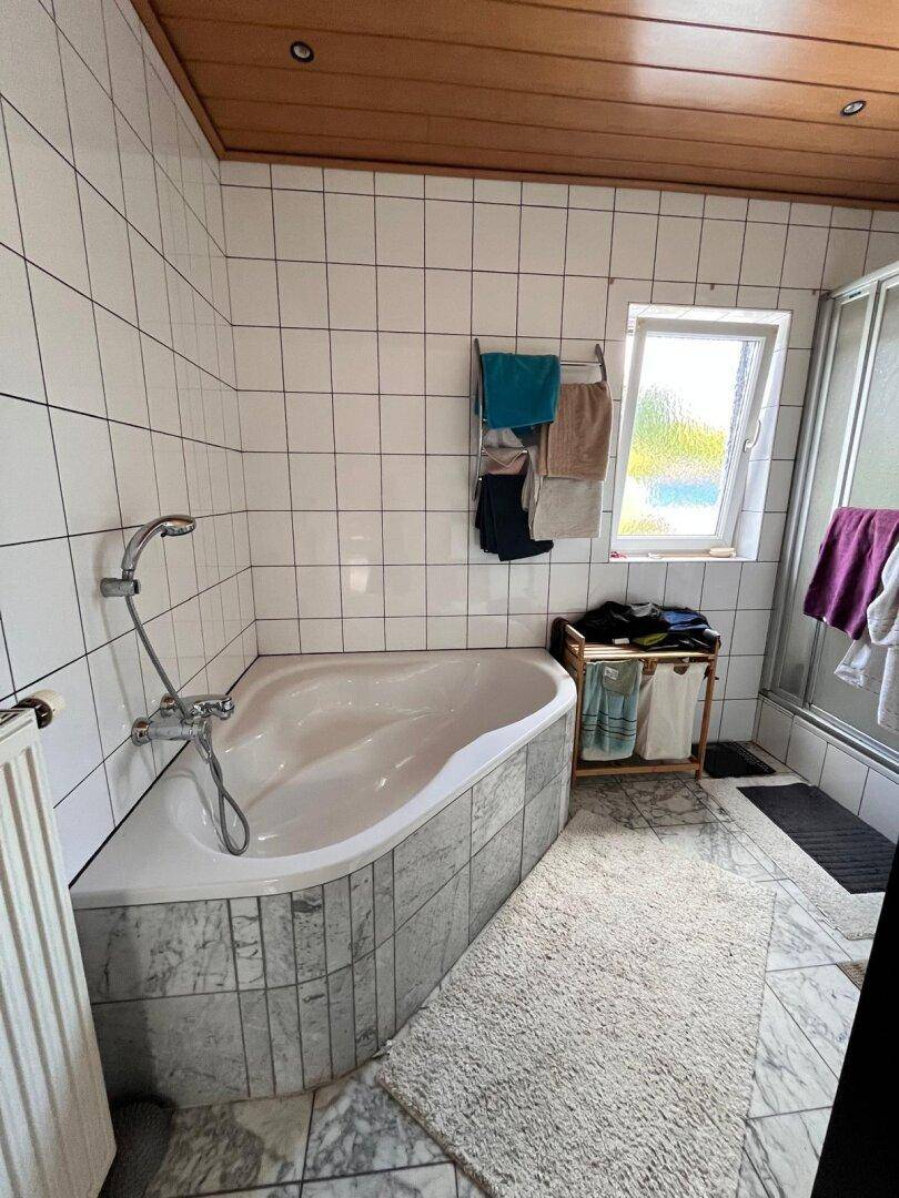 Badezimmer mit Eckbadewanne, weißen Fliesen und Holzdecke, sowie einem Fenster.