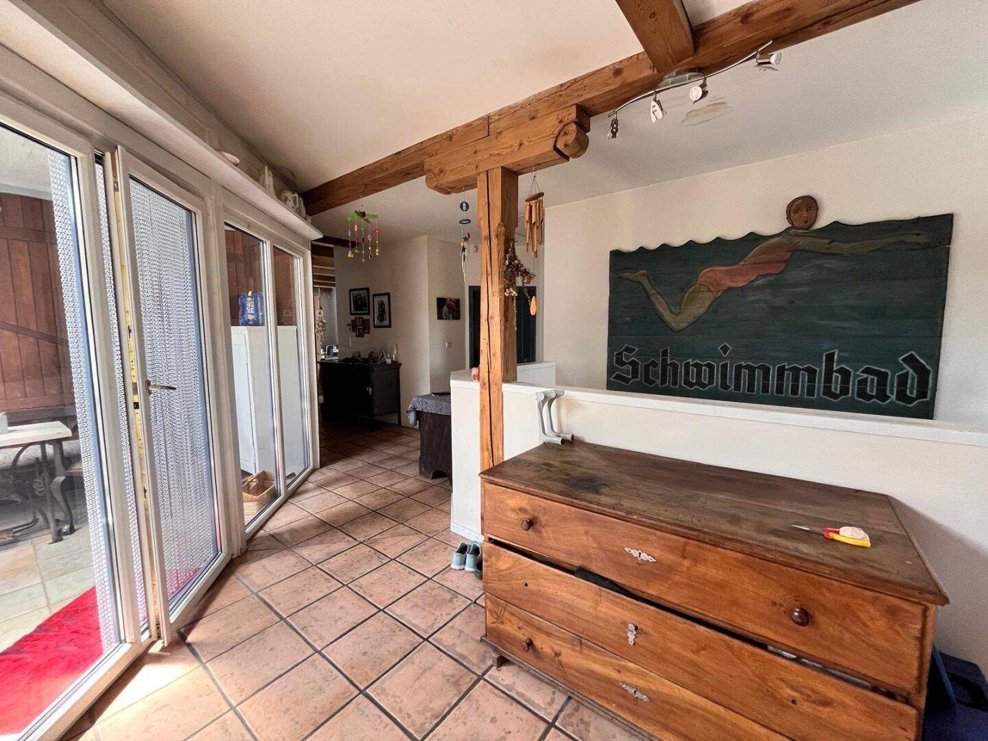 Heller Flur mit Terrakottafliesen, Holzbalken und großen Fenstern, die zu einem Außenbereich führen.
