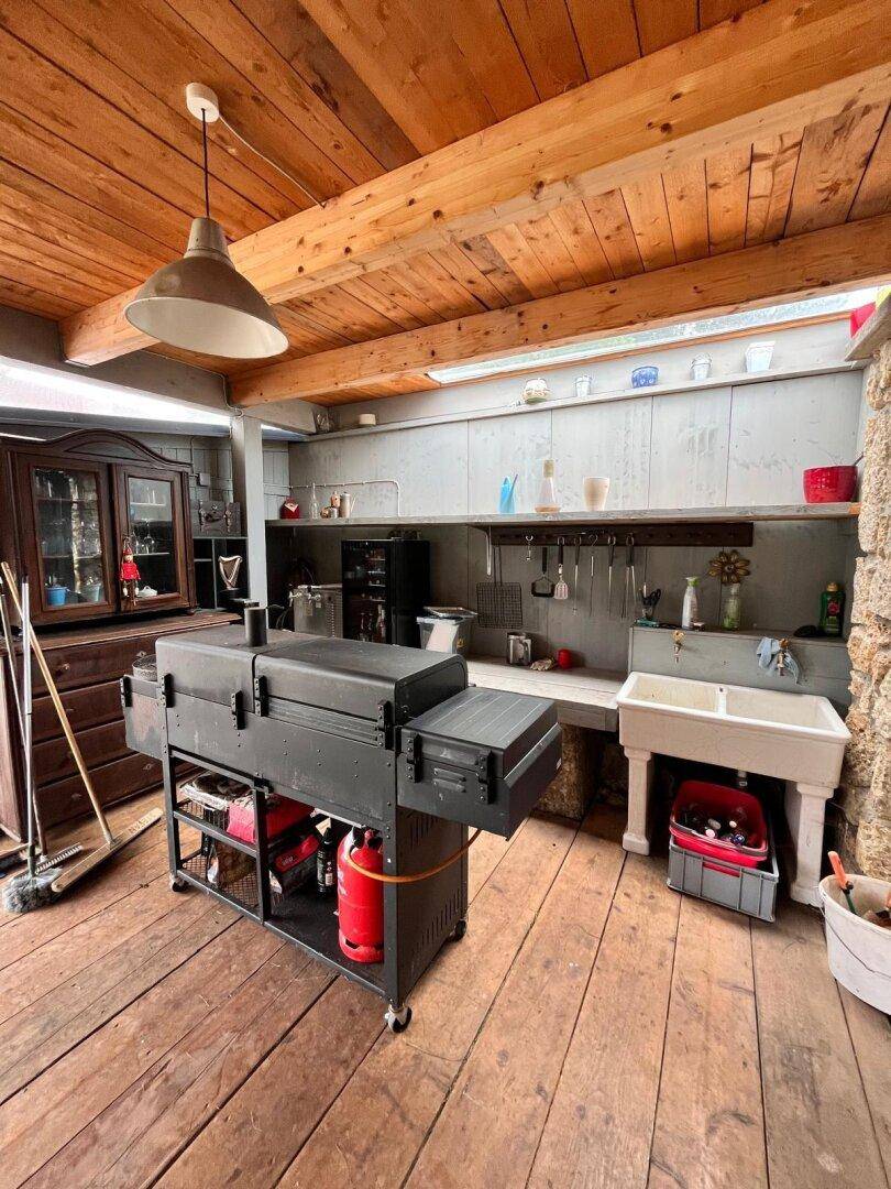 Gut ausgestattete Außenküche unter einer Holzbalkendecke mit Grill und Spülbereich.