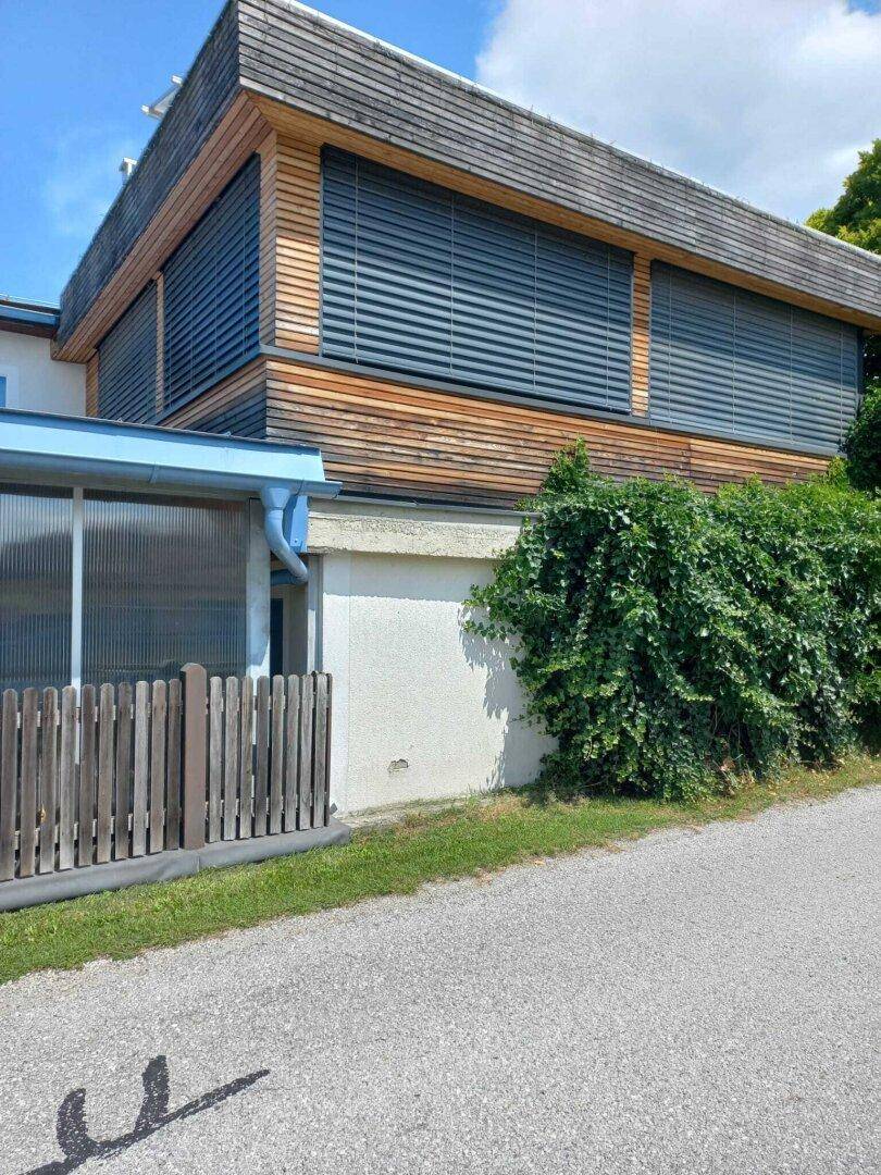 Seitenansicht eines modernen Gebäudes mit Holzfassade, großen Fenstern und Außenjalousien.