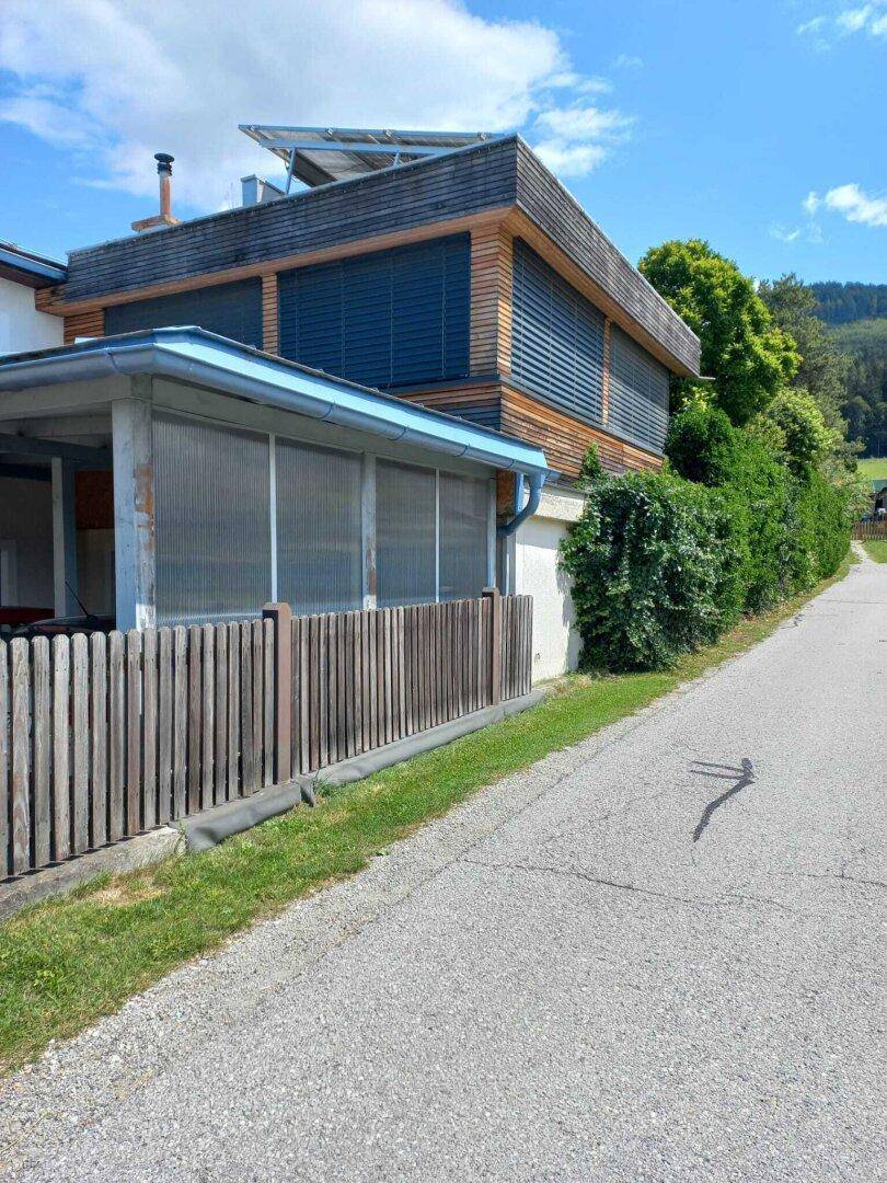 Seitenansicht des Hauses mit Holzverkleidung, Jalousien und einem Zaun entlang der Straße.