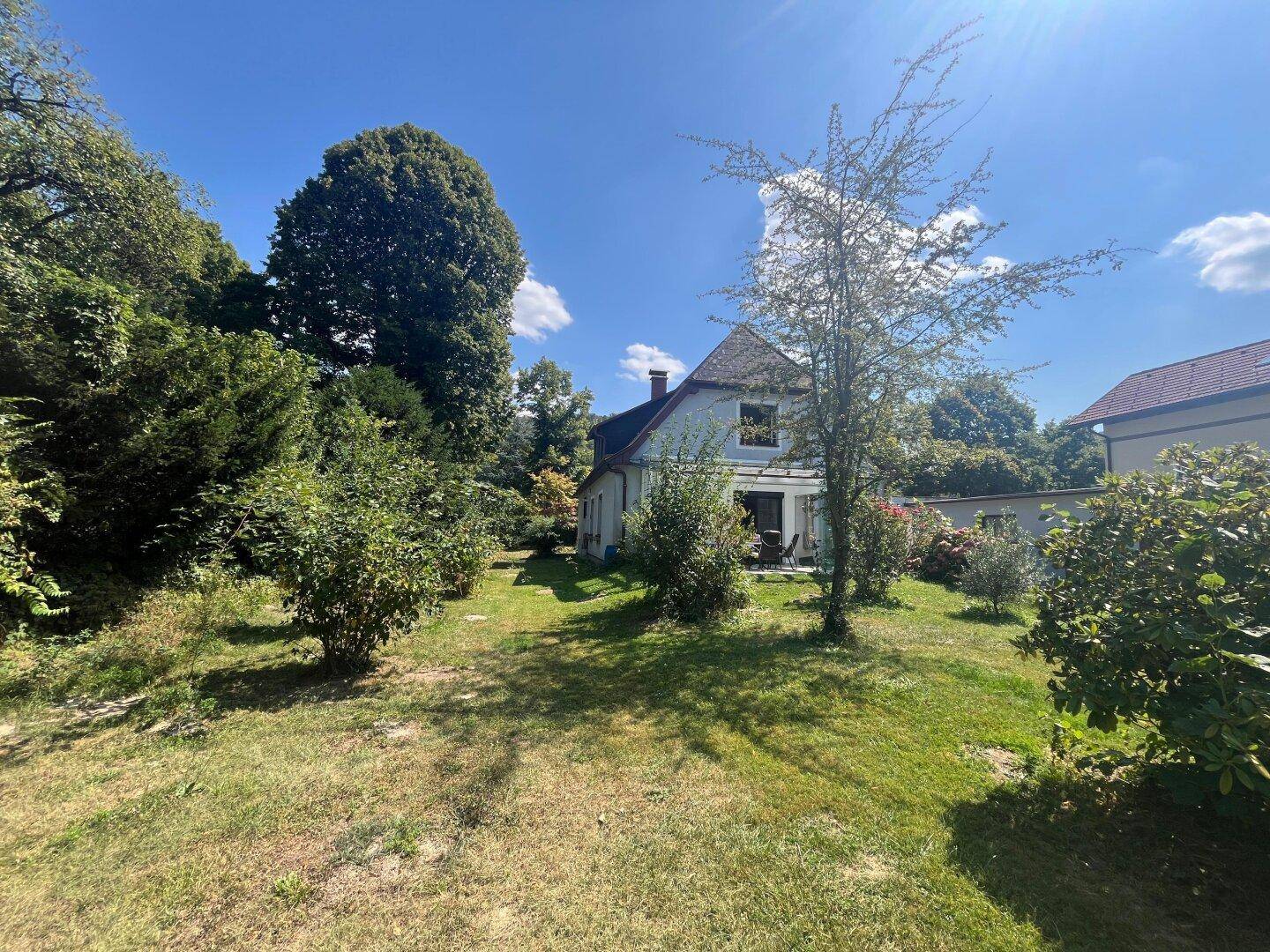 Weitläufiger Garten mit Bäumen und Sträuchern, das Haus ist im Hintergrund sichtbar.
