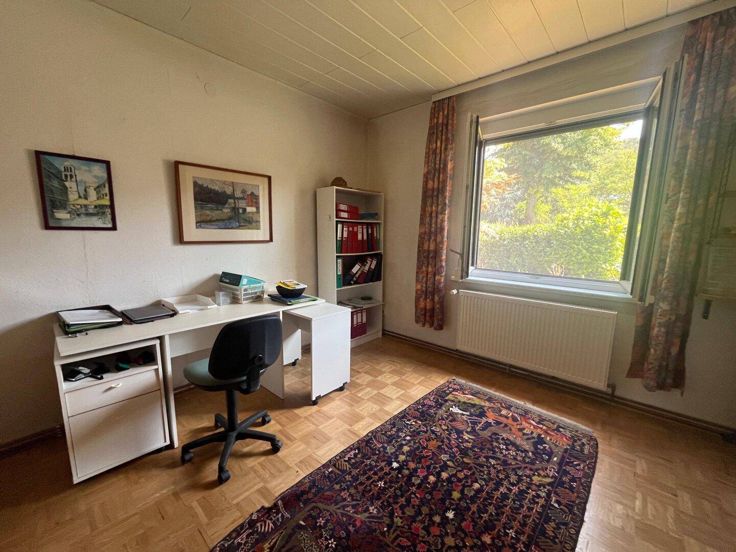 Arbeitszimmer mit Schreibtisch, Regal und Parkettboden, bietet einen ruhigen Blick ins Grüne.