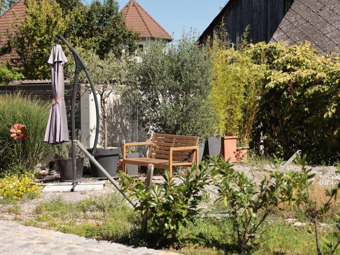 Entspannender Gartenbereich mit einer Holzbank, verschiedenen Topfpflanzen und einem Sonnenschirm.