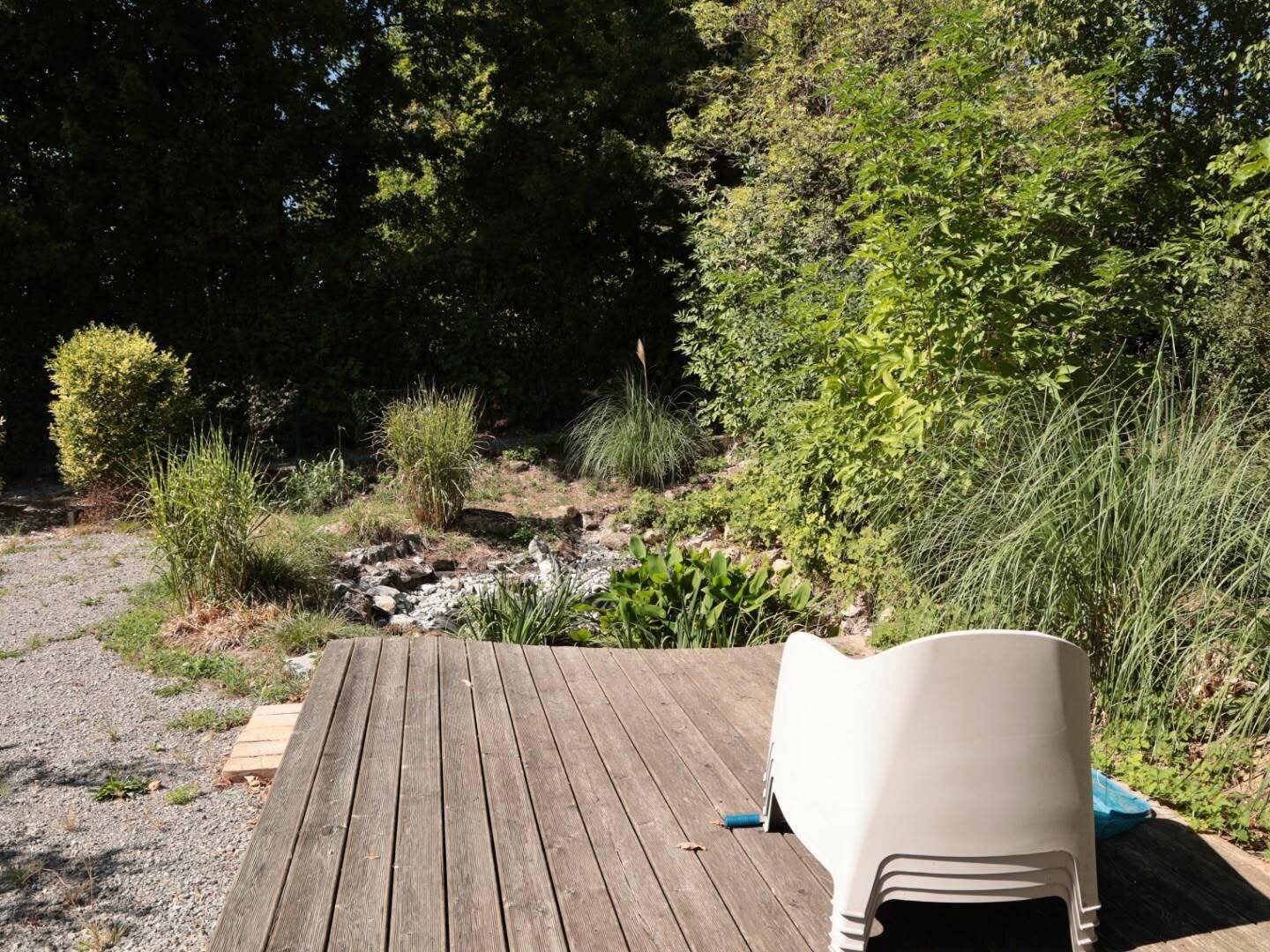 Holzterrasse mit gestapelten Gartenstühlen, die auf einen naturbelassenen Gartenbereich blickt.