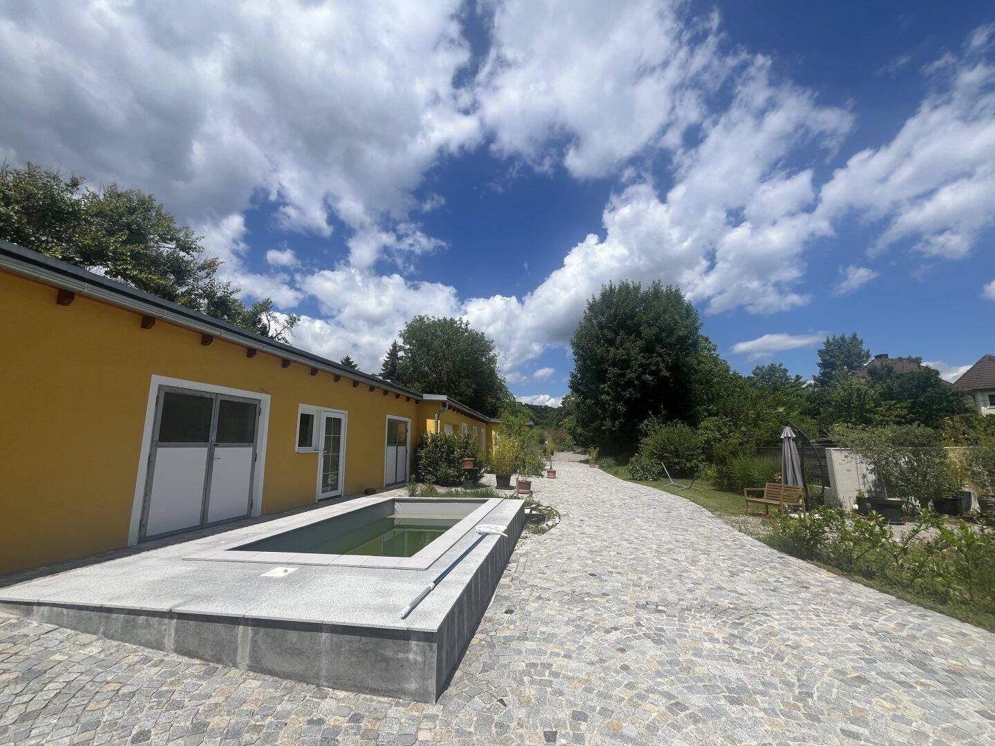 Seitenansicht des Hauses mit gepflastertem Weg, kleinem Wasserbecken und viel Grün.
