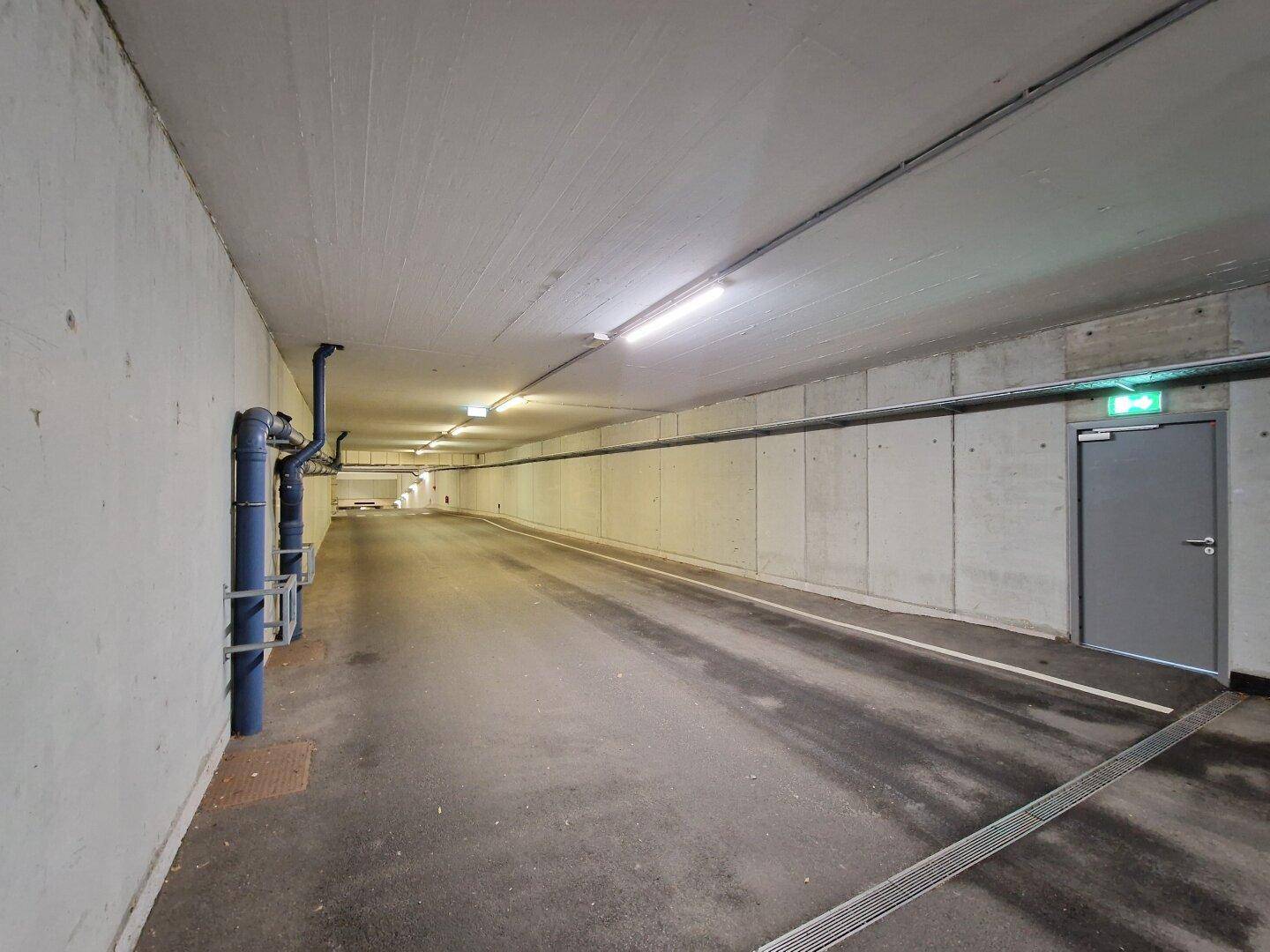 Heller Tiefgaragenzugang mit Betonwänden, blauen Rohren und einer Brandschutztür am Ende des Ganges.