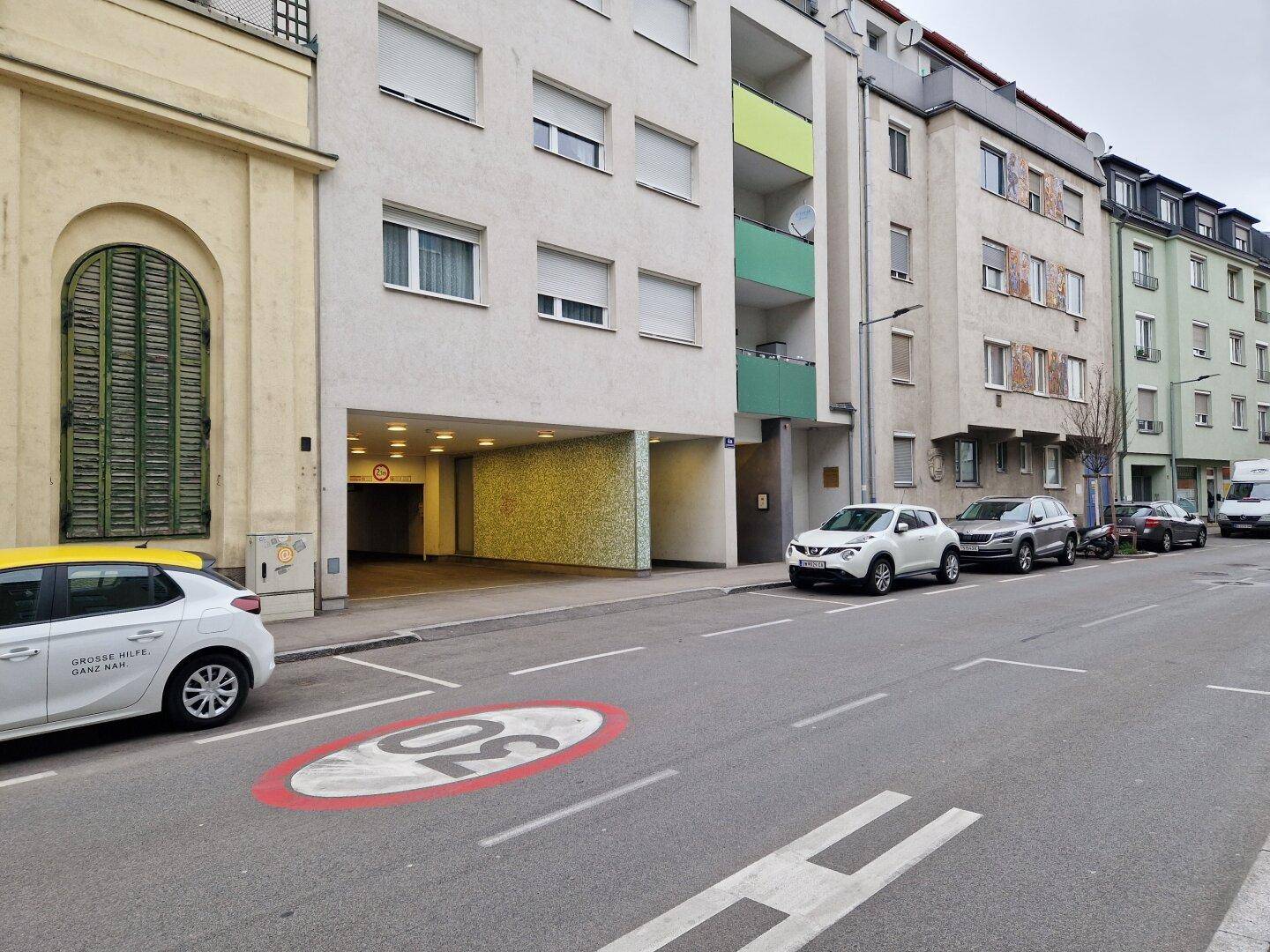 Straßenansicht eines Wohngebäudes mit Tiefgarageneinfahrt und parkenden Autos am Straßenrand.