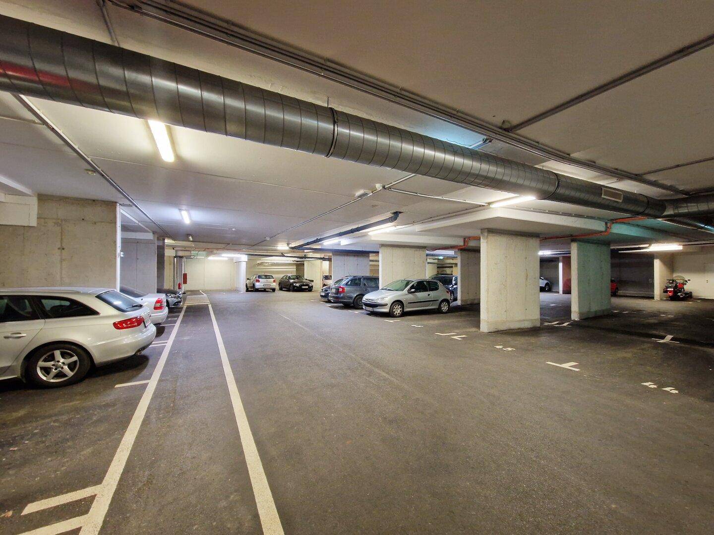 Geräumige Tiefgarage mit vielen Stellplätzen, Betonpfeilern und sichtbaren Lüftungsrohren an der Decke.