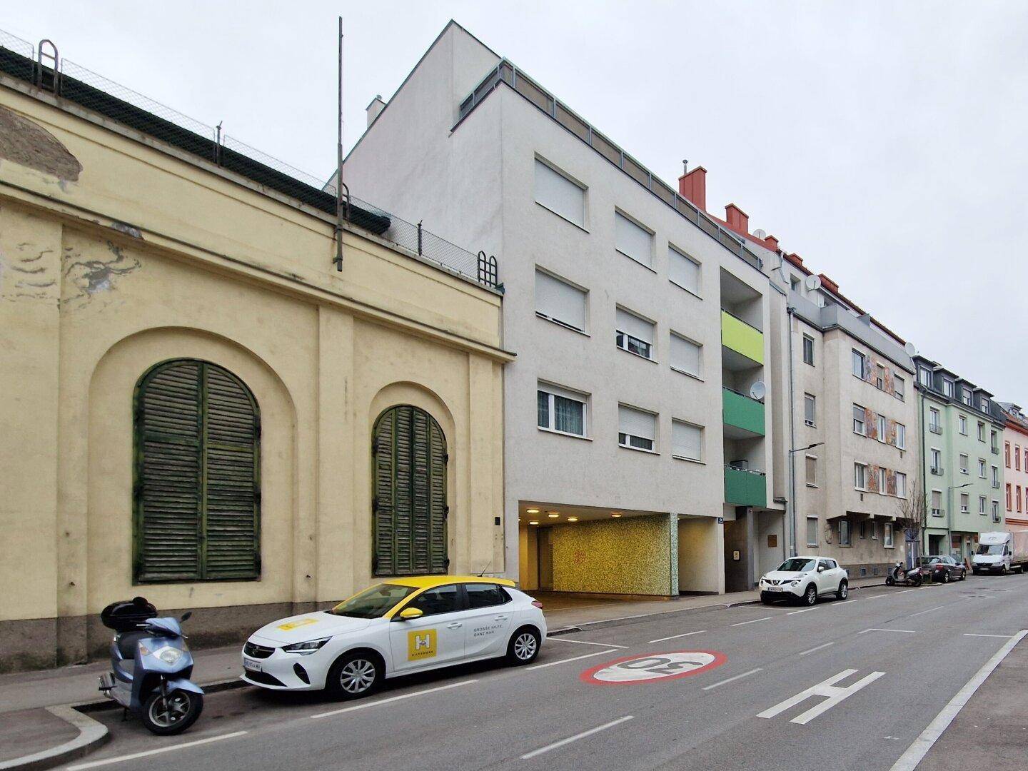 Wohngebäude an einer Straße mit Tiefgarageneinfahrt, umgeben von weiteren Gebäuden und parkenden Autos.