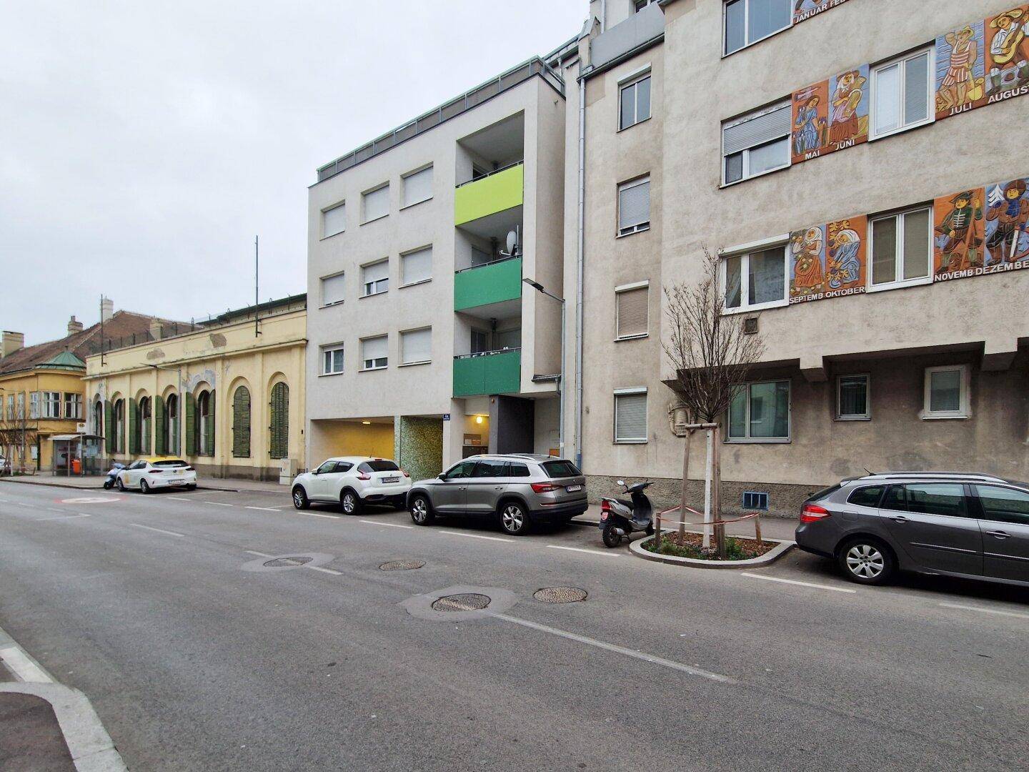 Stadtansicht mit einem modernen Wohngebäude, angrenzenden Altbauten und parkenden Fahrzeugen am Straßenrand.