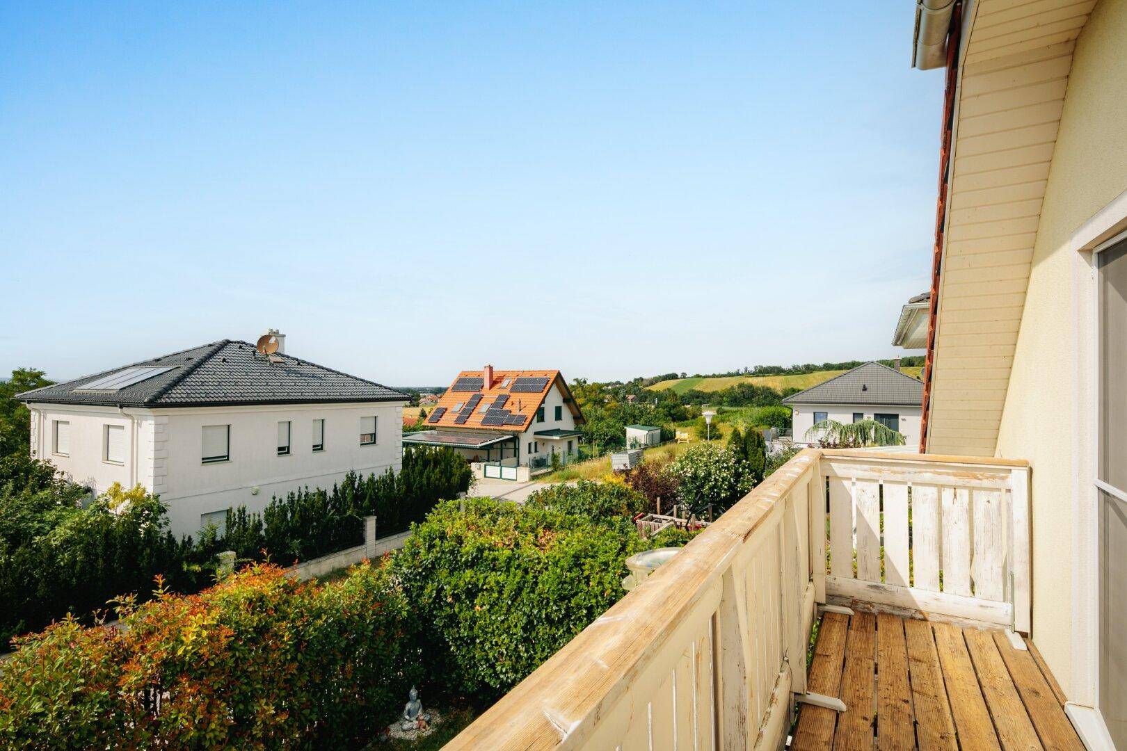 Blick vom Balkon auf die grüne Umgebung und die Dächer der Nachbarhäuser unter blauem Himmel.