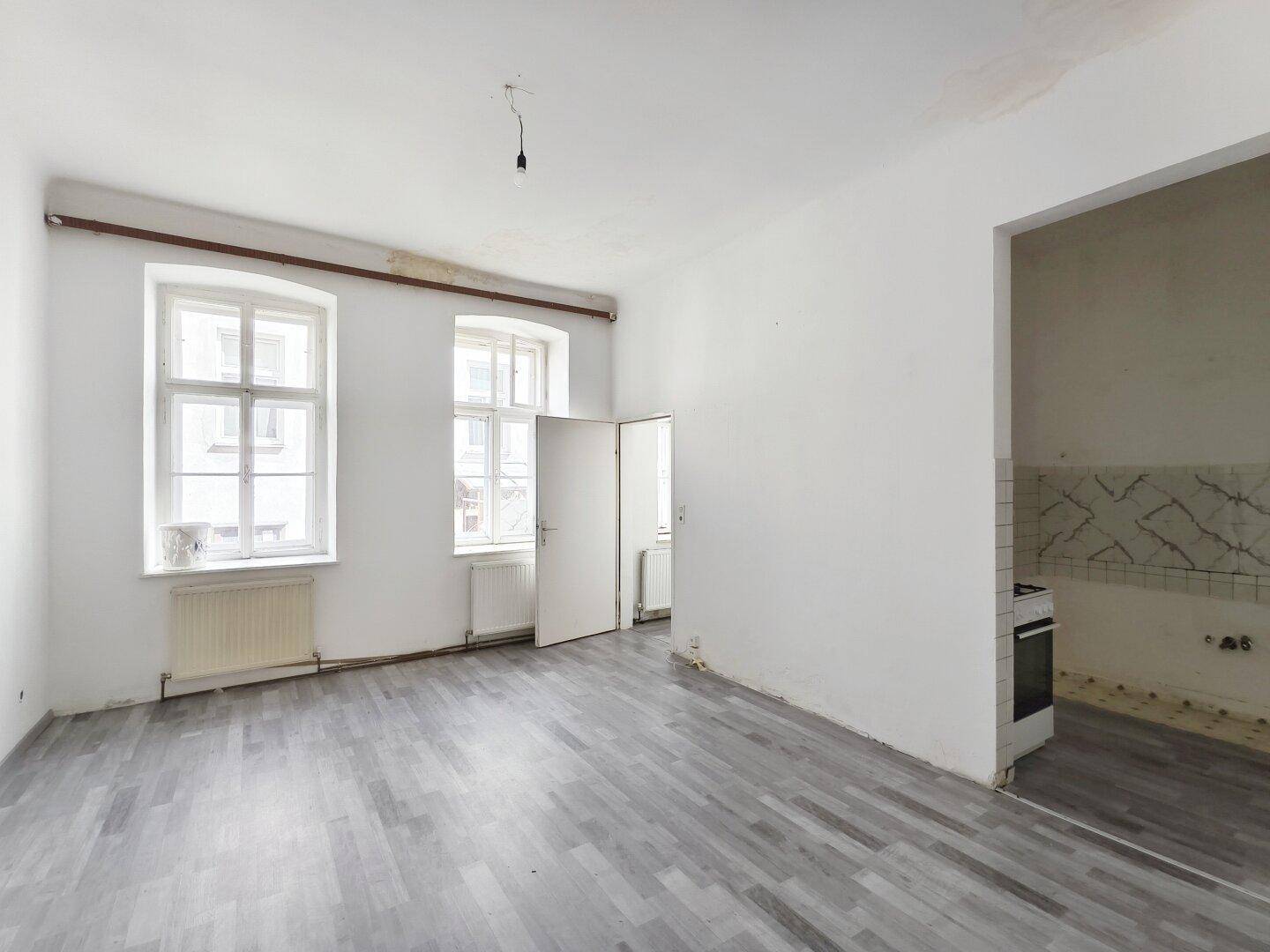 Geräumiges Zimmer mit grauem Laminatboden und Blick in einen angrenzenden Küchenbereich.
