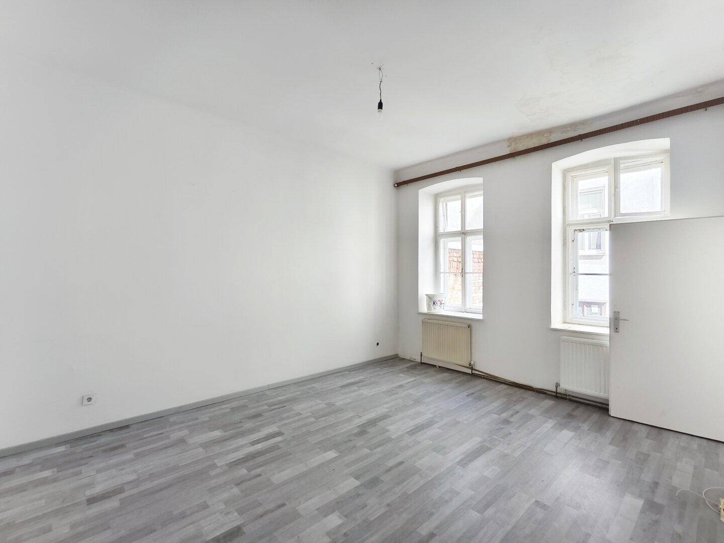Leeres Zimmer mit modernem grauen Laminatboden und zwei großen Fenstern.