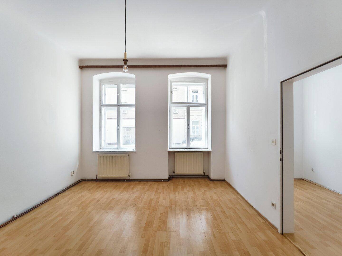 Helles, leeres Zimmer mit Parkettboden und zwei Fenstern, bereit zur individuellen Gestaltung.