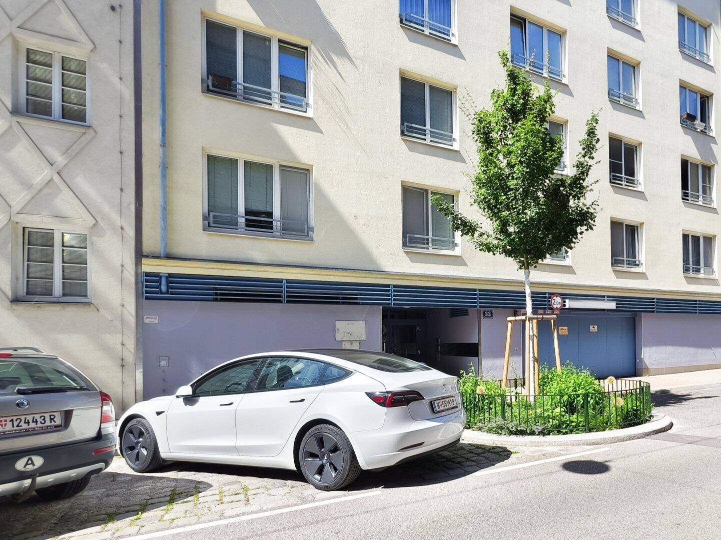 Außenansicht eines Wohngebäudes mit Tiefgarageneinfahrt und einem geparkten weißen Elektrofahrzeug.
