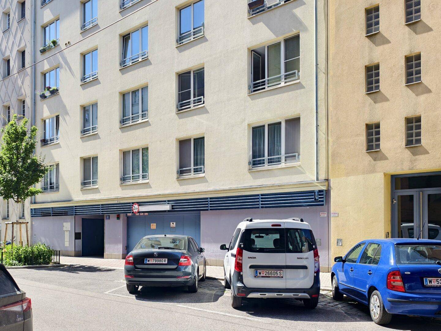 Gesamtansicht der Gebäudefassade mit Tiefgarageneinfahrt und mehreren geparkten Fahrzeugen auf der Straße.