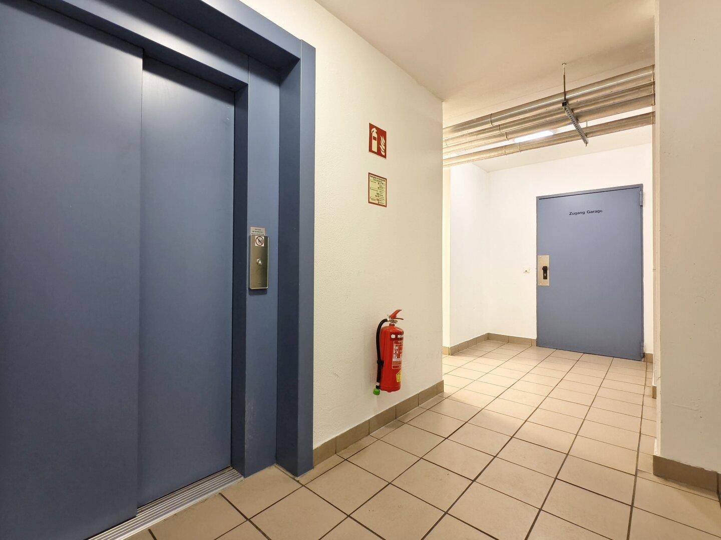 Flur mit Aufzug, Fliesenboden und einer Tür, die den Zugang zur Tiefgarage ermöglicht.
