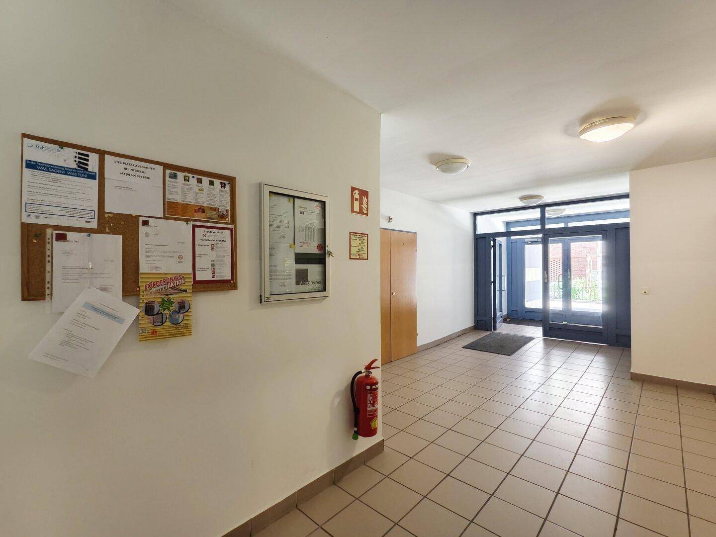 Eingangsbereich des Gebäudes mit Fliesenboden, Informationstafel und Glastüren zum Außenbereich.