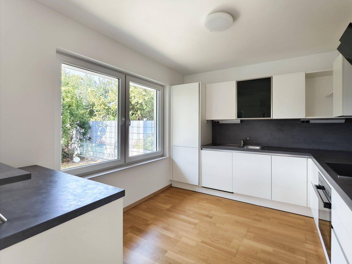 Helle Küche mit modernen weißen Schränken, grauer Arbeitsplatte und Fenster zum Garten.