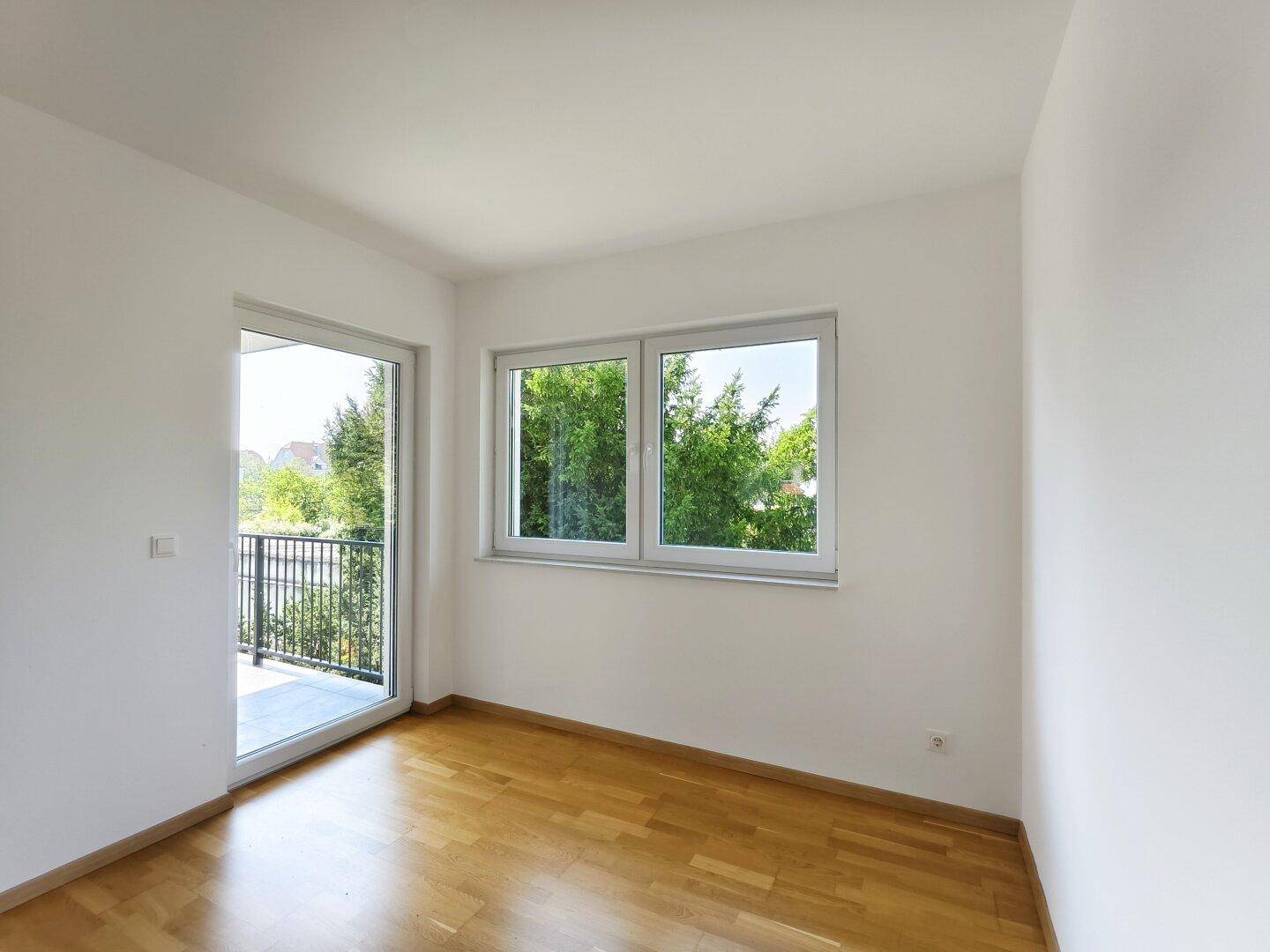 Helles Schlafzimmer mit Holzboden, Fenster und Balkontür, die den Raum mit Licht füllen.