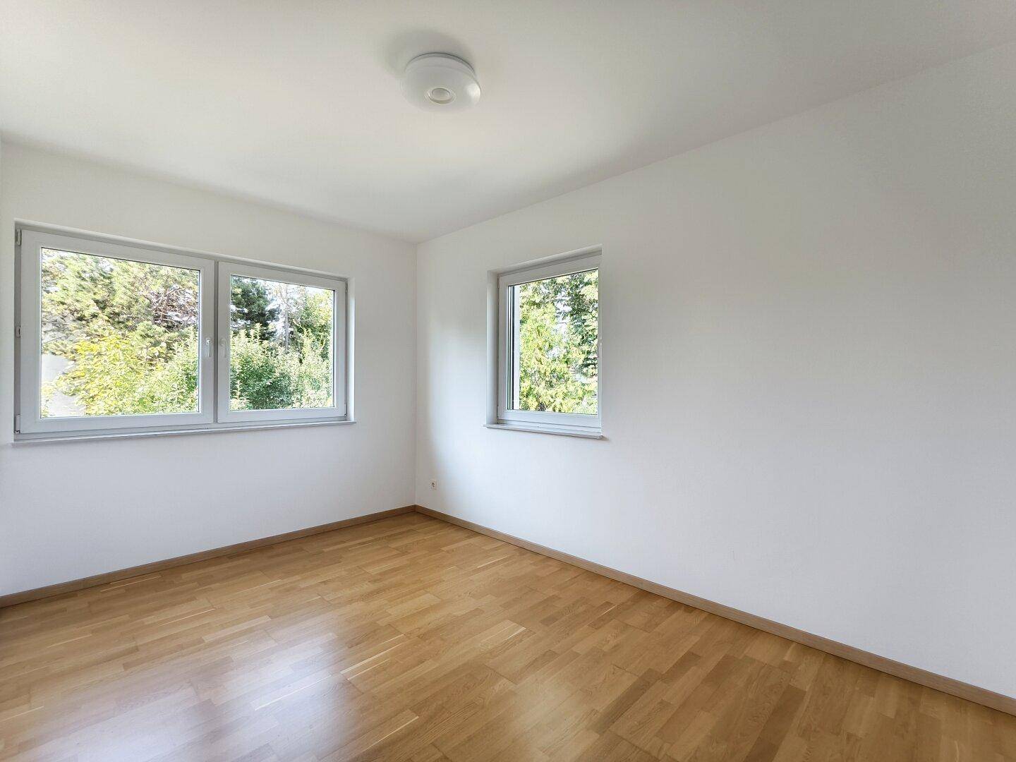 Helles Zimmer mit Parkettboden und mehreren Fenstern, die viel Tageslicht hereinlassen.