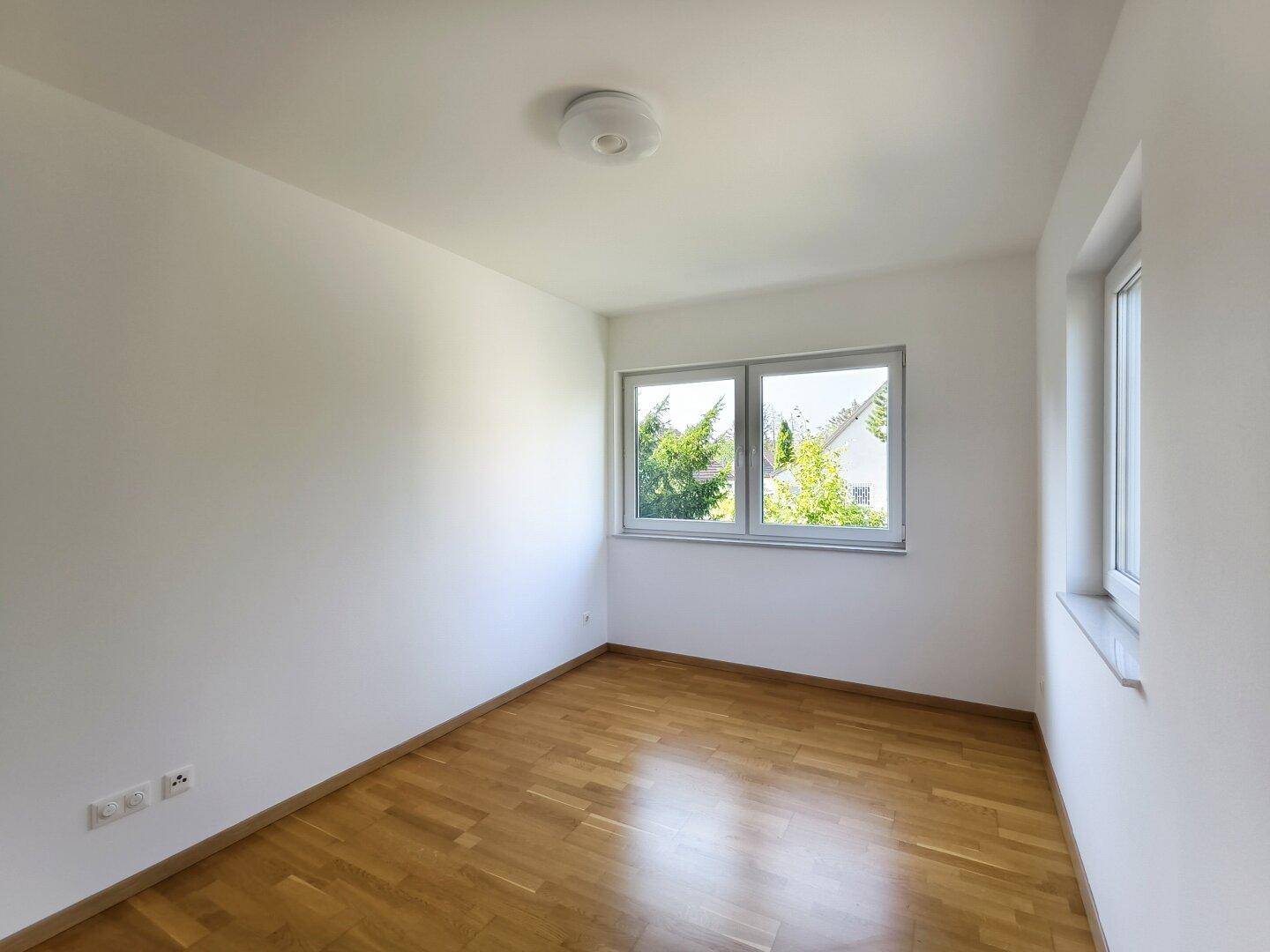 Leeres, helles Zimmer mit Parkettboden und Blick ins Grüne durch die Fenster.