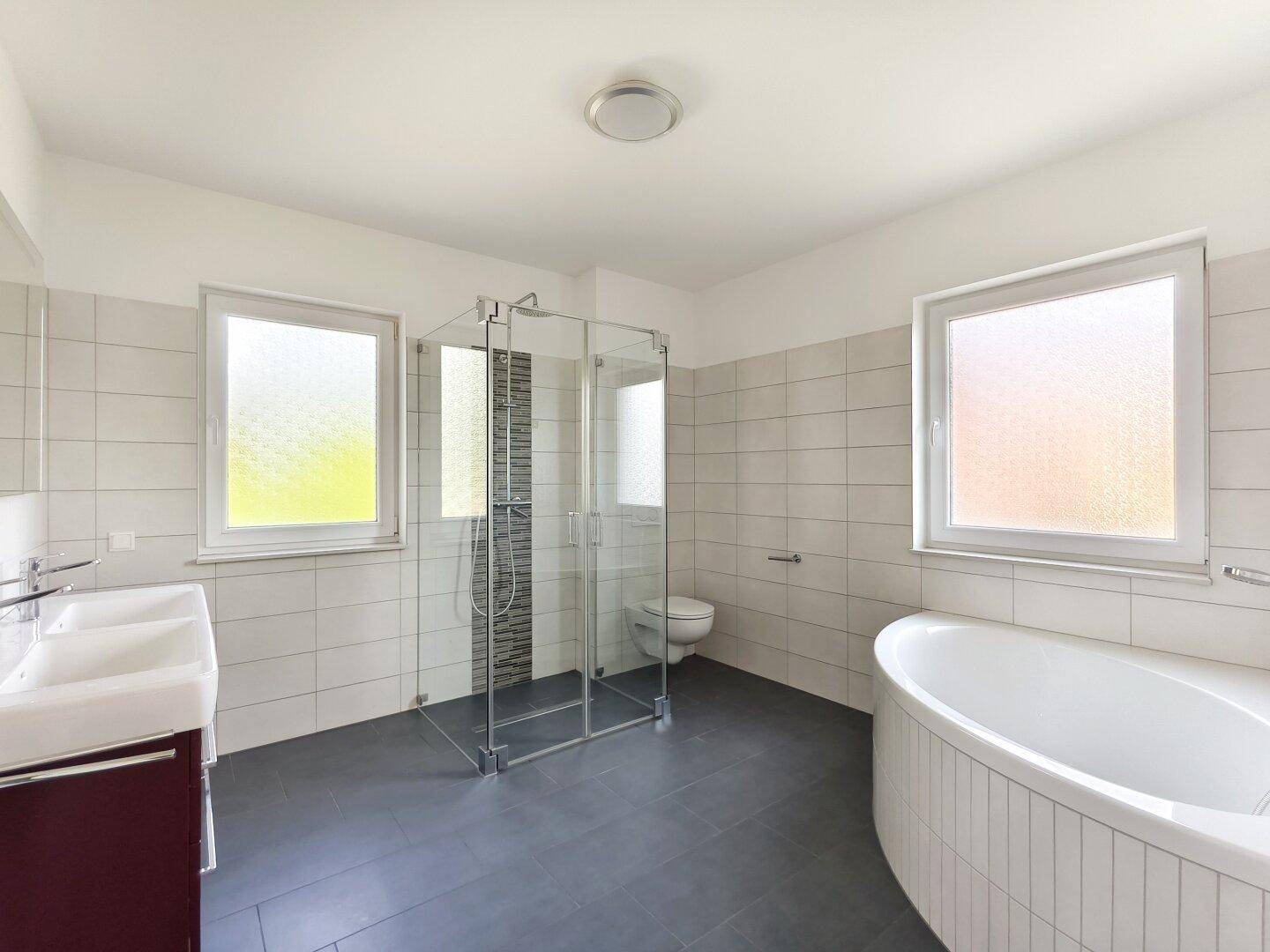 Geräumiges Badezimmer mit Badewanne, großer Dusche und zwei Fenstern für Tageslicht.