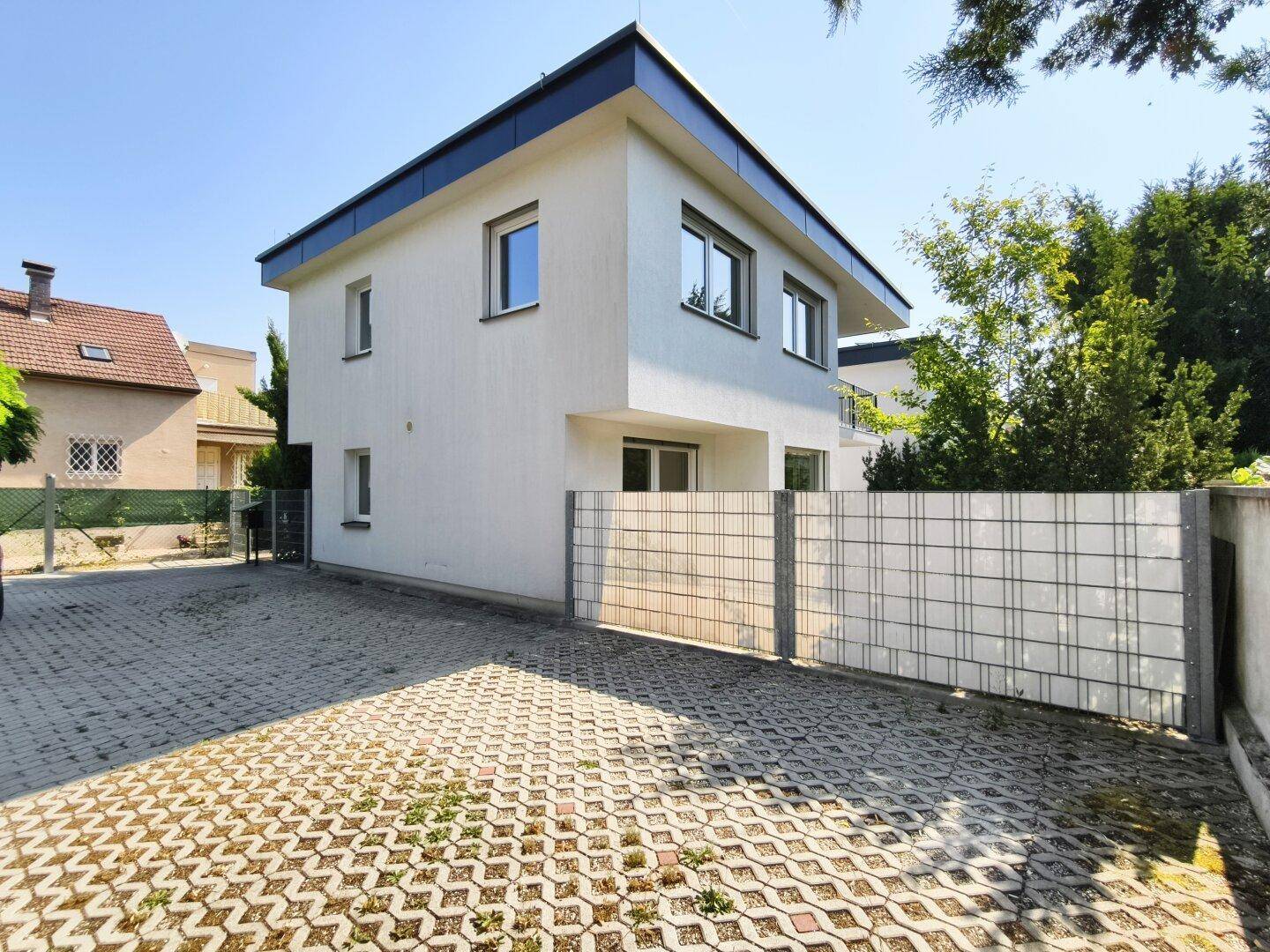 Modernes Wohngebäude mit weißer Fassade und gepflastertem Vorplatz an einem sonnigen Tag.
