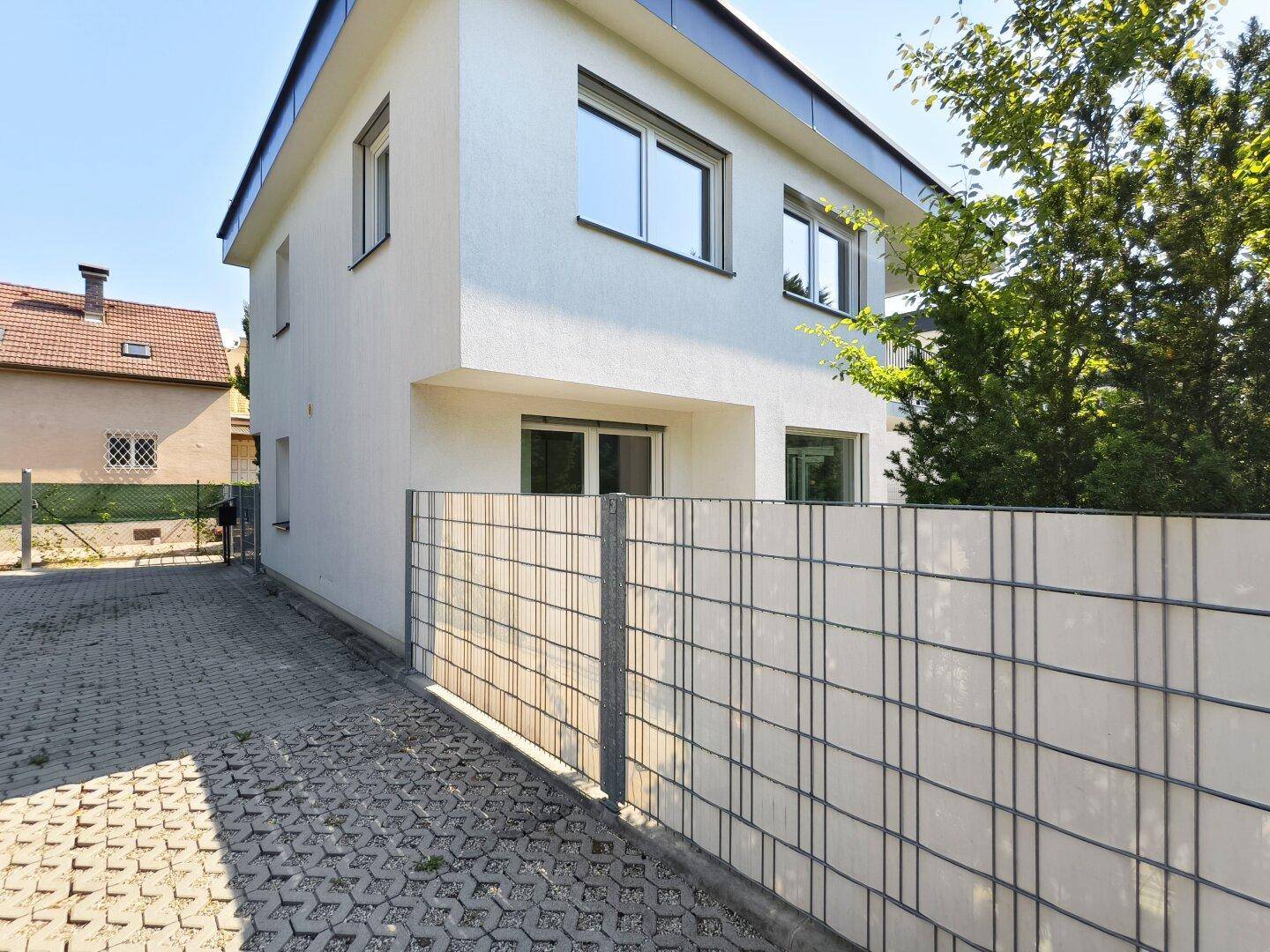 Seitenansicht eines modernen Neubaus mit weißer Fassade und gepflastertem Zugangsbereich.