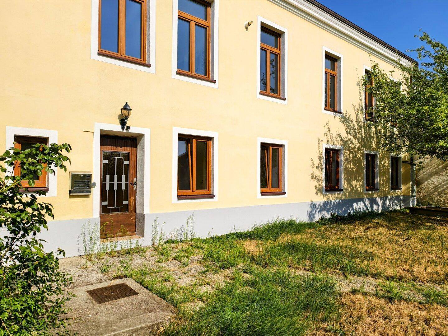 Gelbe Fassade eines Altbaus mit braunen Fenstern und einem ungepflegten Vorgarten.