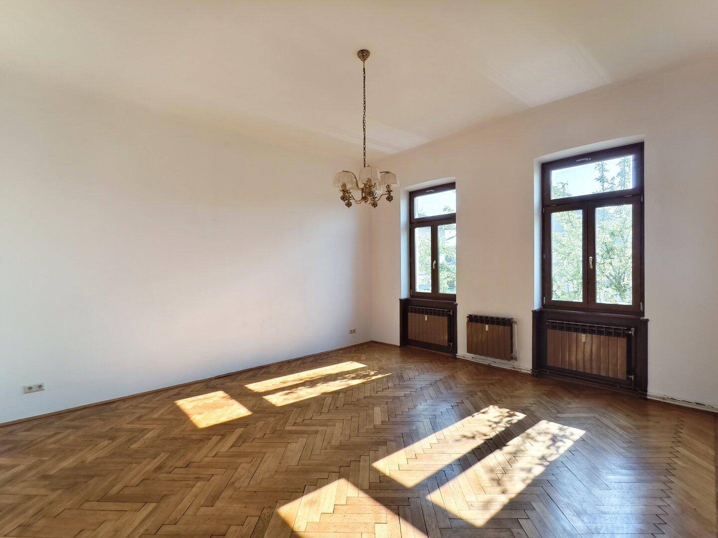 Lichtdurchflutetes Zimmer mit Fischgrätparkett und Kronleuchter, ideal für einen Wohnbereich.