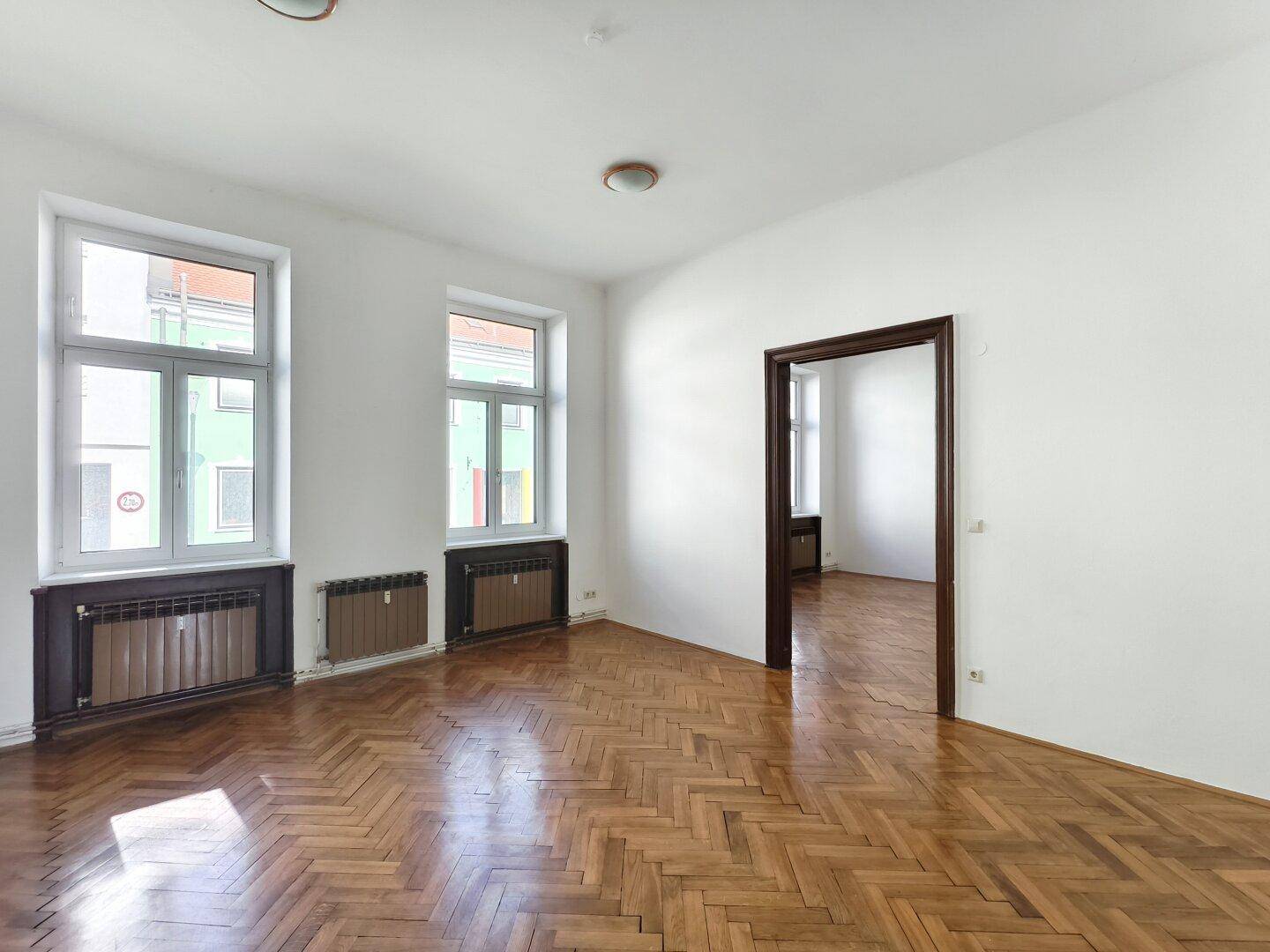 Helles, leeres Zimmer mit Fischgrätparkett und großen Fenstern zur Straße.