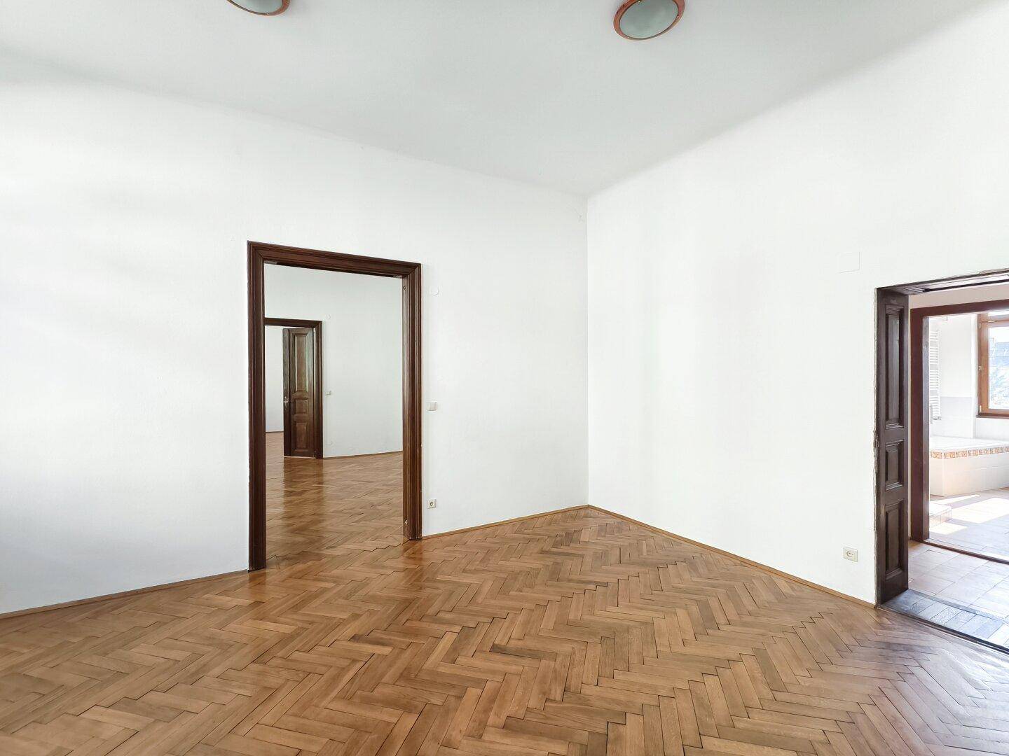 Geräumiges Zimmer mit Fischgrätparkett und hohen Holztüren, die verschiedene Bereiche verbinden.