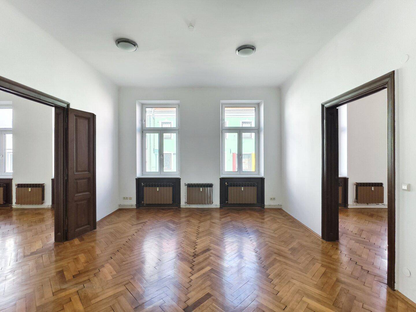 Lichtdurchflutetes Zimmer mit Fischgrätparkett und zwei großen Fenstern.