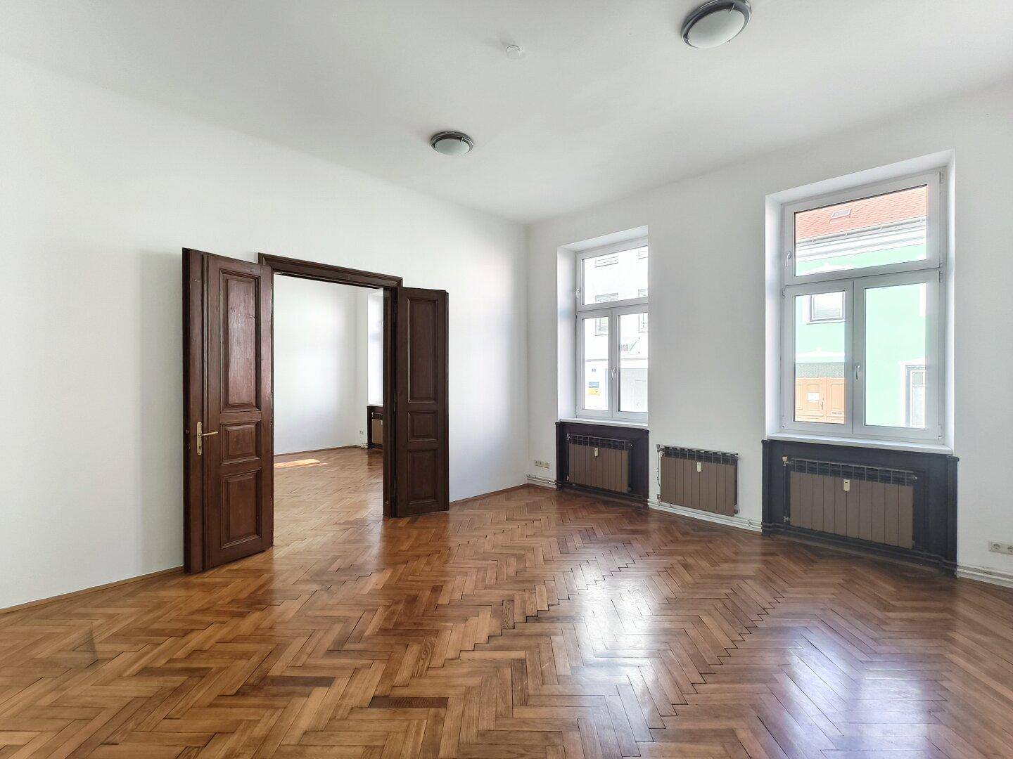 Helles Zimmer mit Fischgrätparkett und hohen Fenstern, die viel Tageslicht hereinlassen.