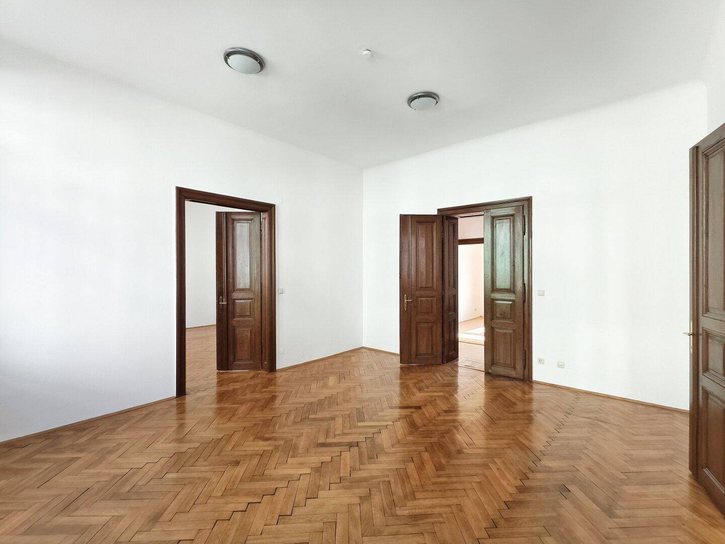 Geräumiges Zimmer mit Fischgrätparkett und hohen Holztüren, die eine offene Raumgestaltung ermöglichen.