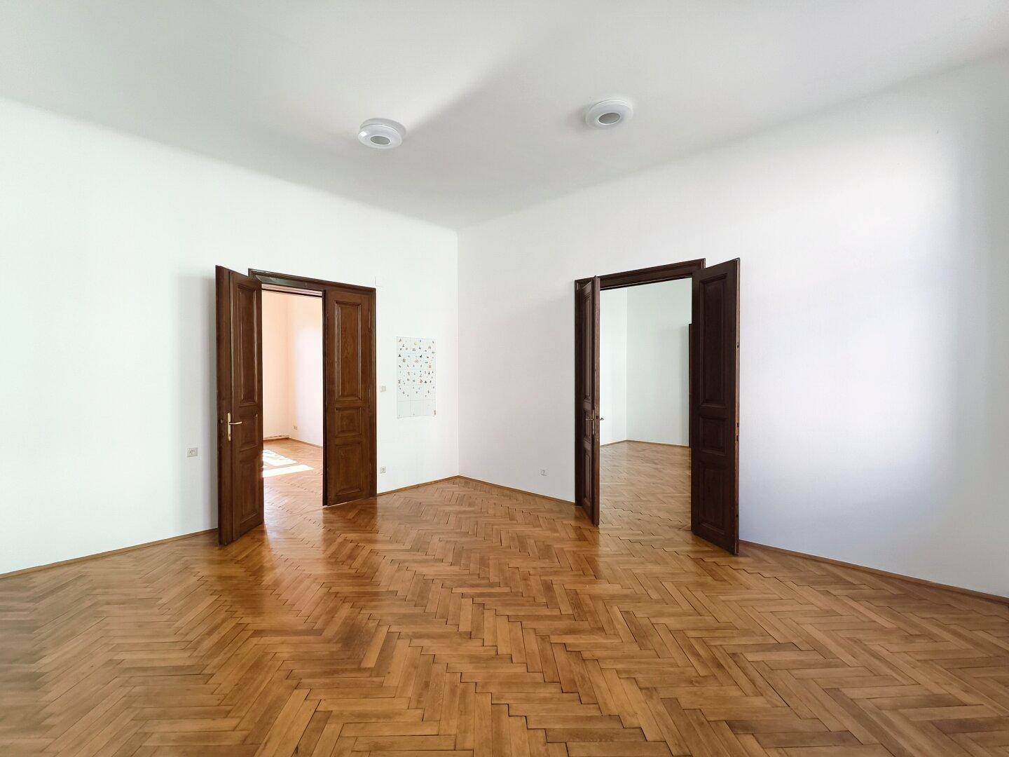 Großes Zimmer mit Fischgrätparkett und mehreren offenen Holztüren, die eine flexible Nutzung bieten.