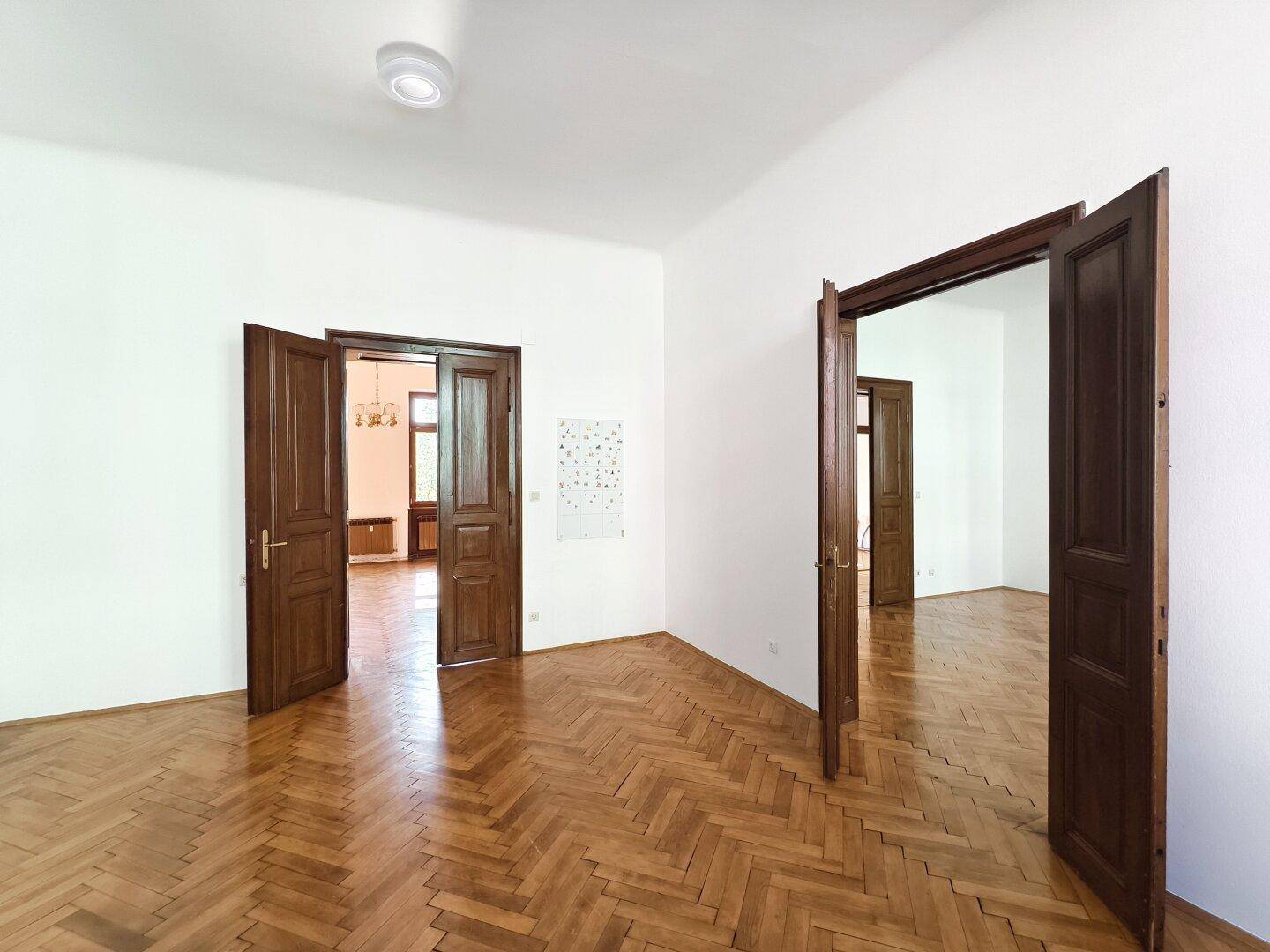 Geräumiges Zimmer mit Fischgrätparkett und hohen Holztüren, die zu angrenzenden Räumen führen.