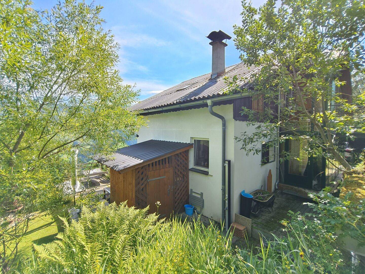 Seitenansicht des Hauses mit einem kleinen Holzschuppen, umgeben von üppigem Grün.