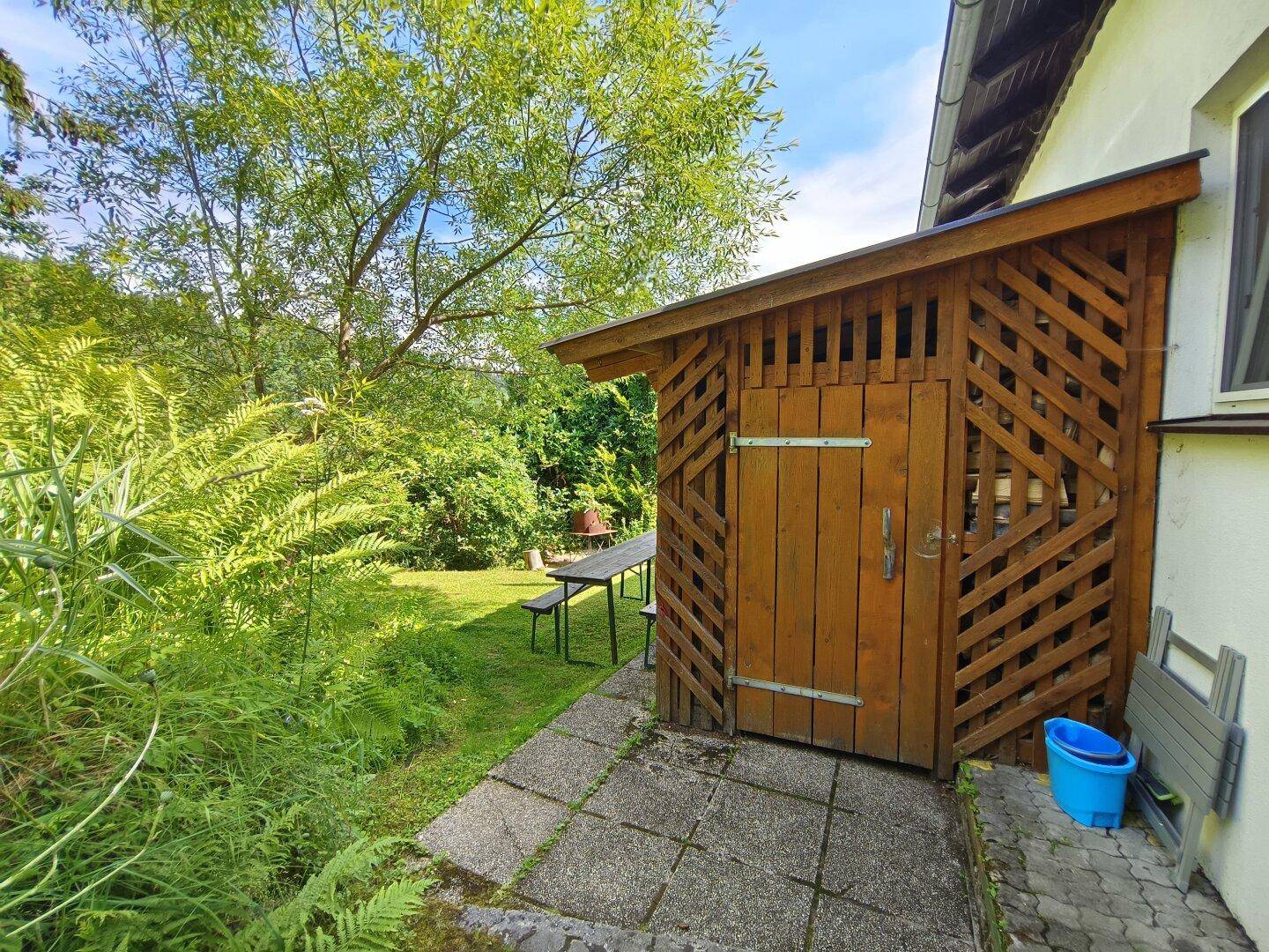 Kleiner Holzschuppen im Gartenbereich neben dem Haus, umgeben von üppigem Grün.