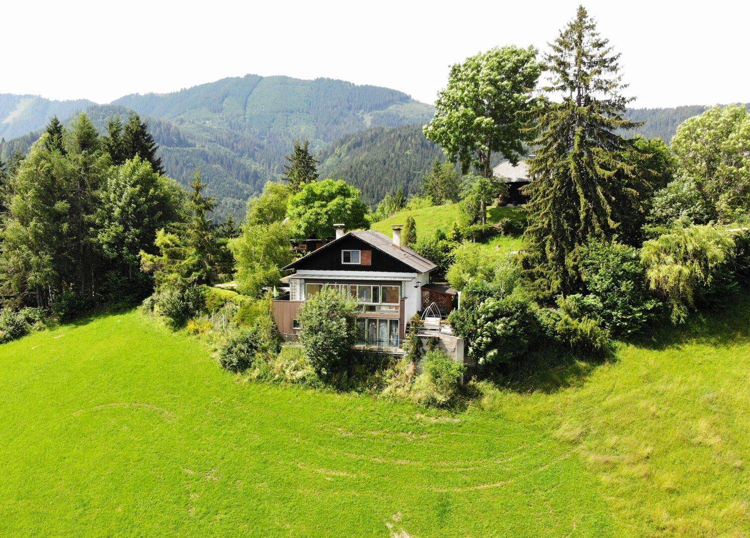 Luftaufnahme eines Hauses auf einem grünen Hügel, umgeben von Bäumen und Bergen.