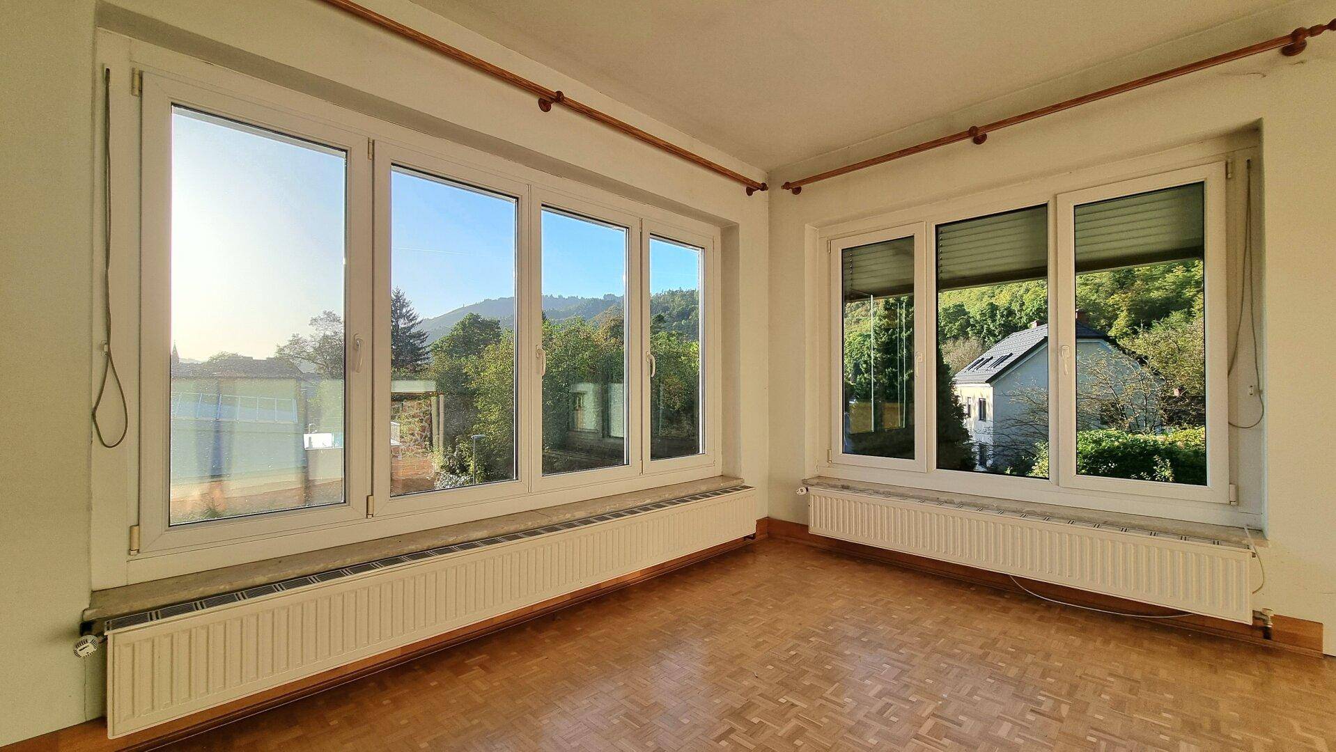 Leerer Raum mit großen Fenstern, Parkettboden und Blick ins Grüne.