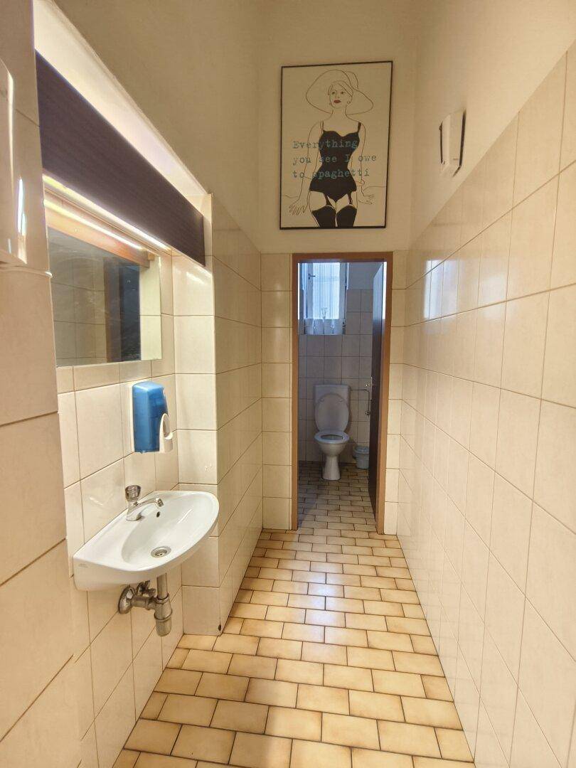 Gefliester Toilettenbereich mit Waschbecken, Spiegel und einer Tür, die zu einer weiteren Toilette führt.