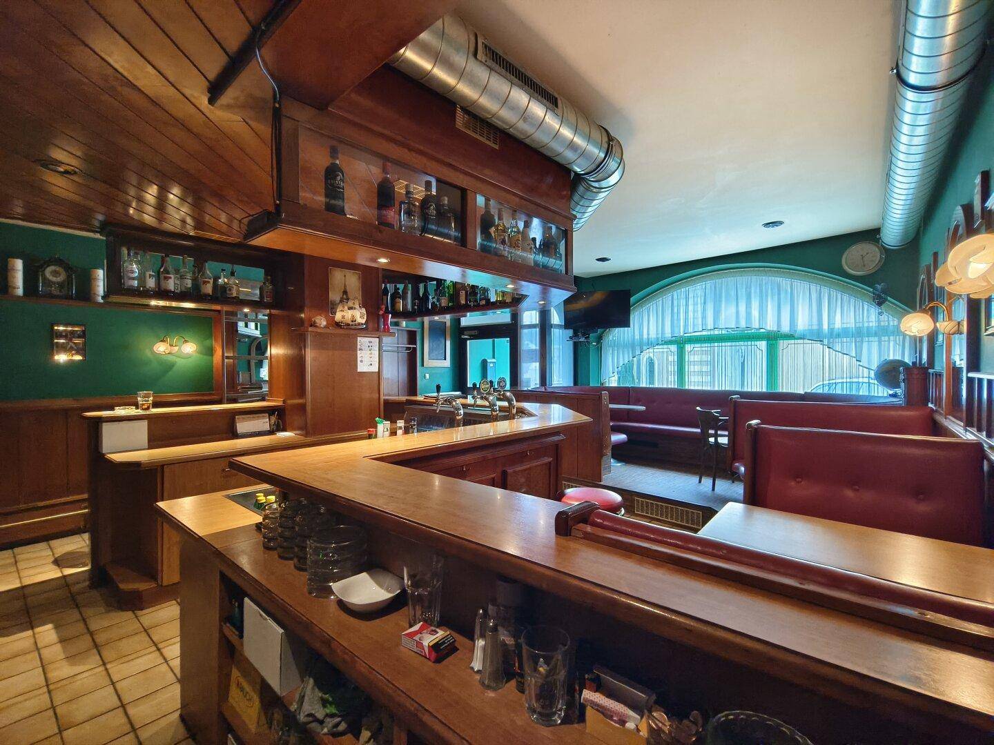 Bar mit Holztheke, Flaschenregalen und Sitzgelegenheiten, beleuchtet durch Fenster mit Vorhängen.