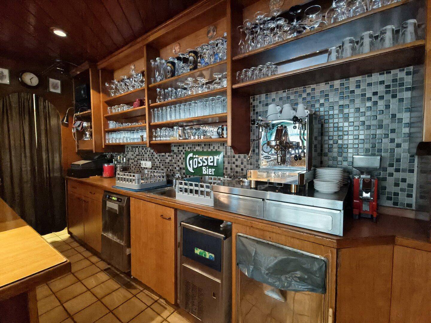 Thekenbereich mit Kaffeemaschine, Gläserregalen und Fliesenrückwand, bereit für den Service.