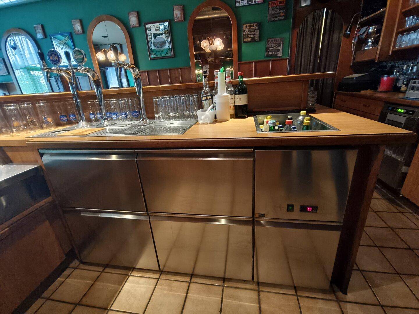 Barfront mit Edelstahl-Kühlschränken, Zapfanlage und Holztheke, bereit für Getränke.