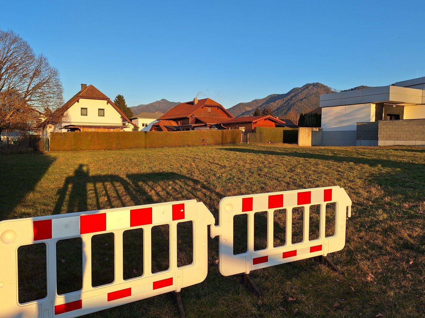 Baugrundstück mit Absperrungen im Vordergrund, umgeben von Wohnhäusern und Berglandschaft.