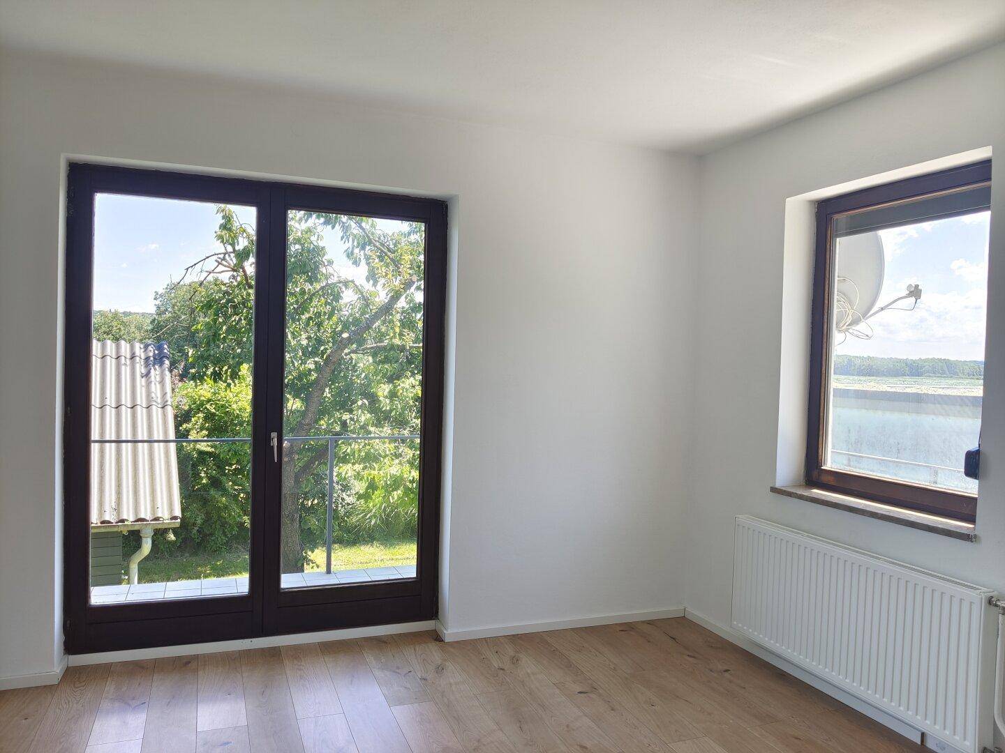 Helles, leeres Zimmer mit Holzboden, weißen Wänden und Zugang zu einem französischen Balkon.